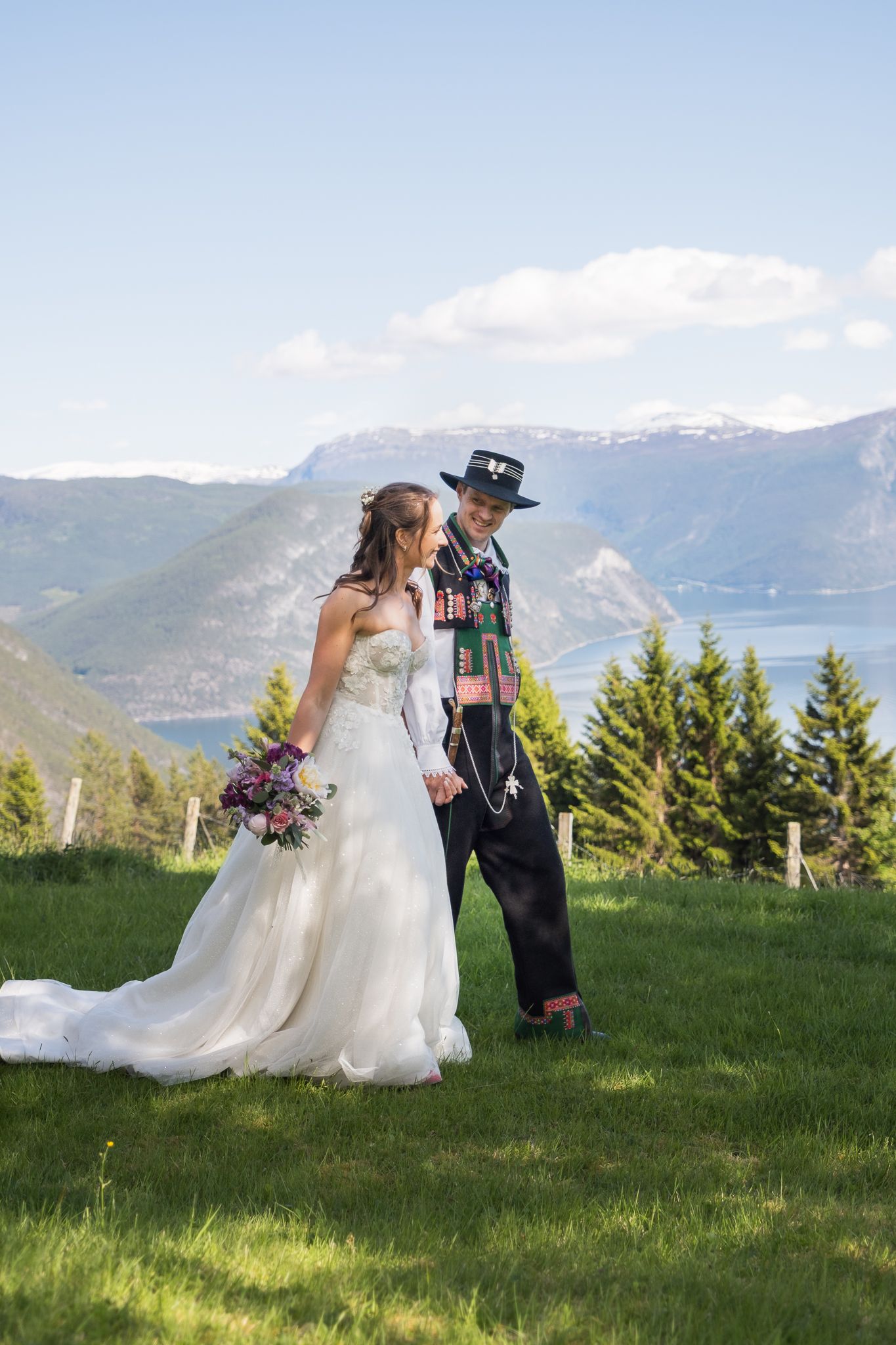 Bride in white gown and groom in traditional Norwegian bunad hold hands on a green hillside with fjord and mountains behind.