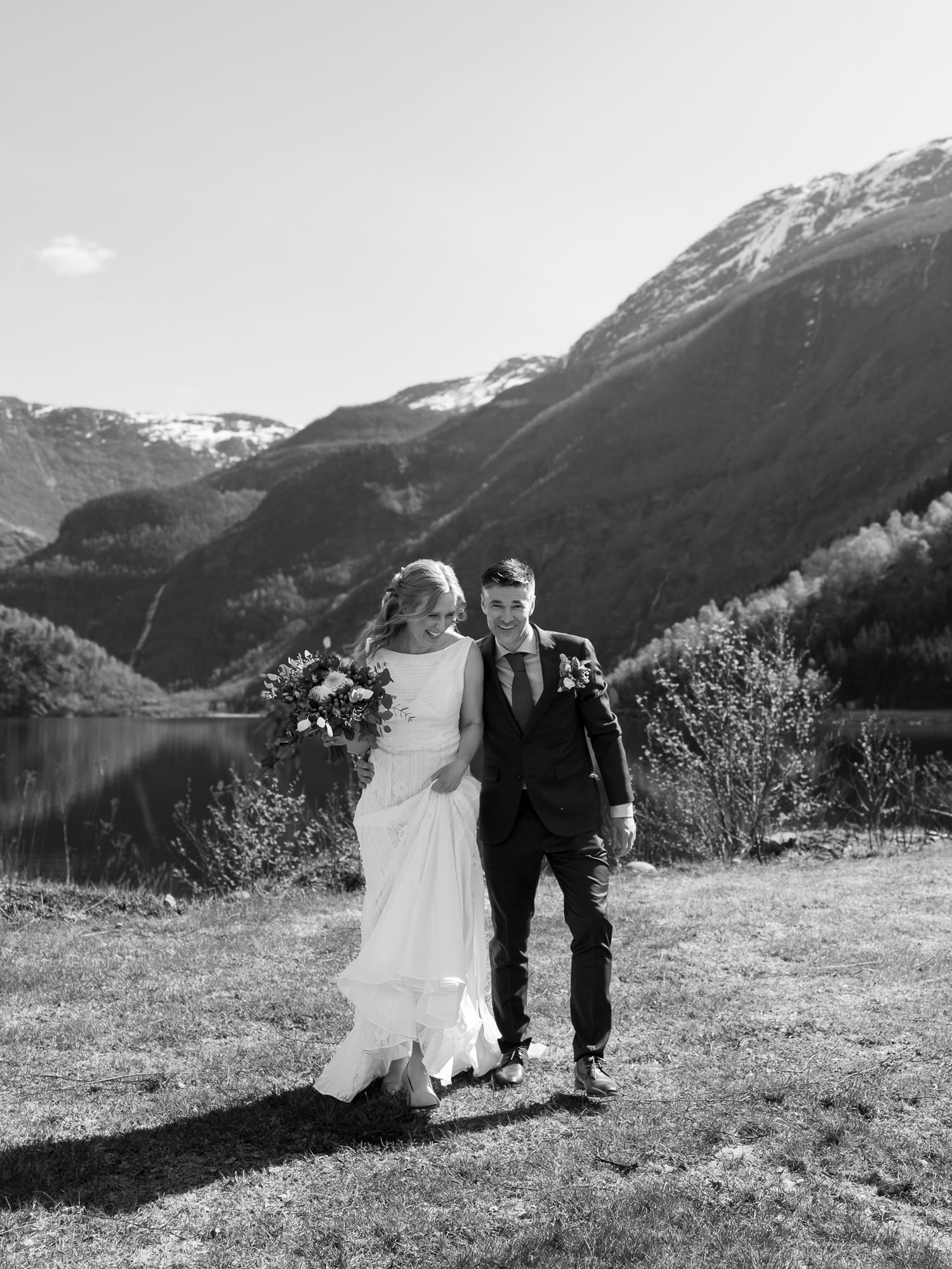 Bride and groom walking by a fjord with dramatic mountain scenery in a black and white wedding portrait.