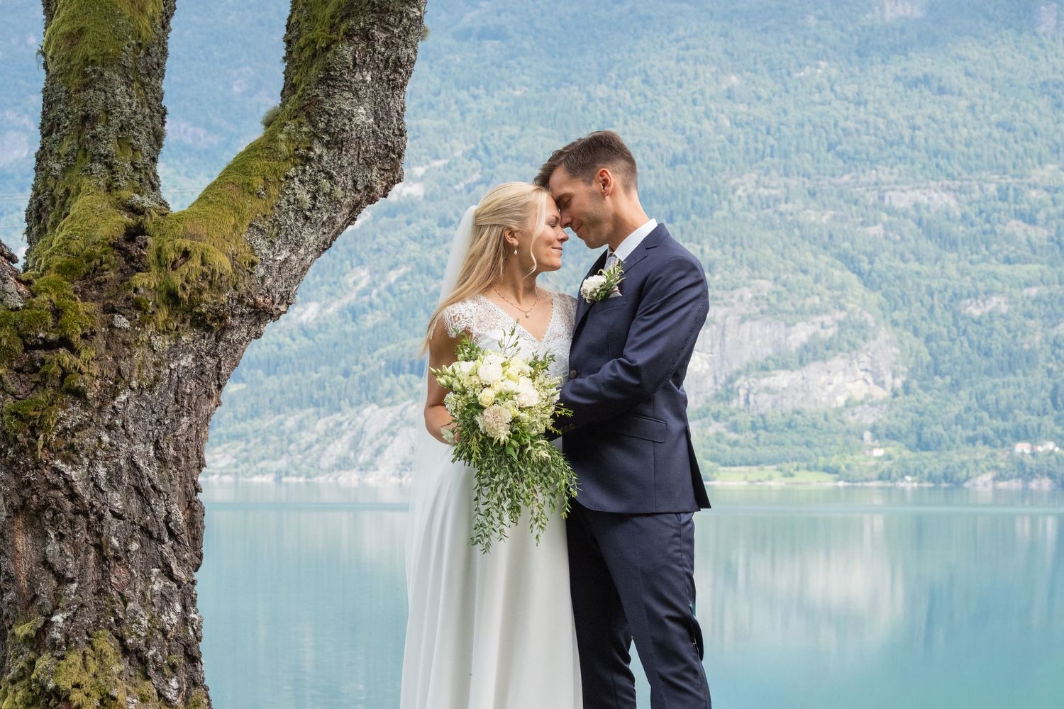 Bride and groom embrace tenderly under mossy tree with scenic fjord and mountains behind them.