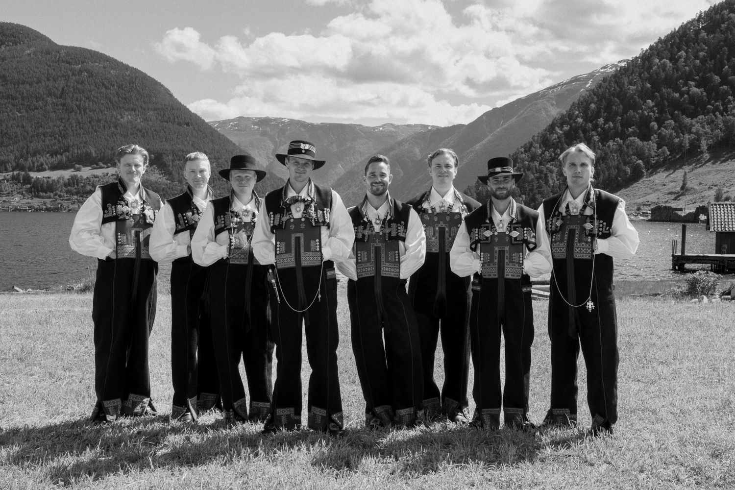 Eight men in traditional Norwegian bunad folk costumes posing in a line by a scenic mountain fjord.