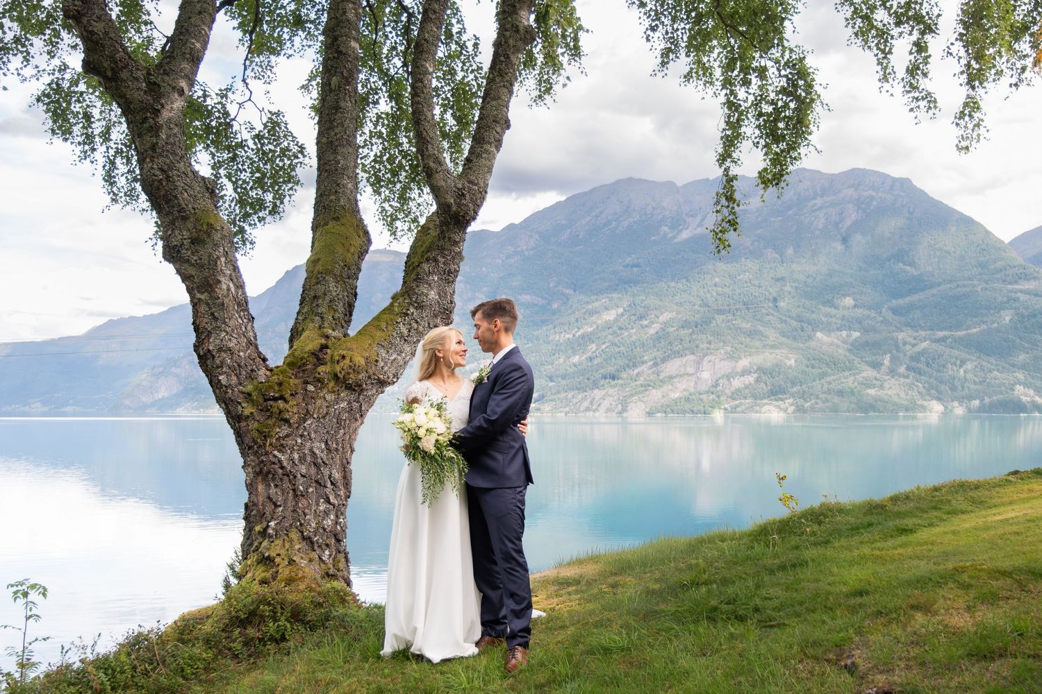 Bride and groom share romantic moment under birch tree with stunning fjord and mountains backdrop.