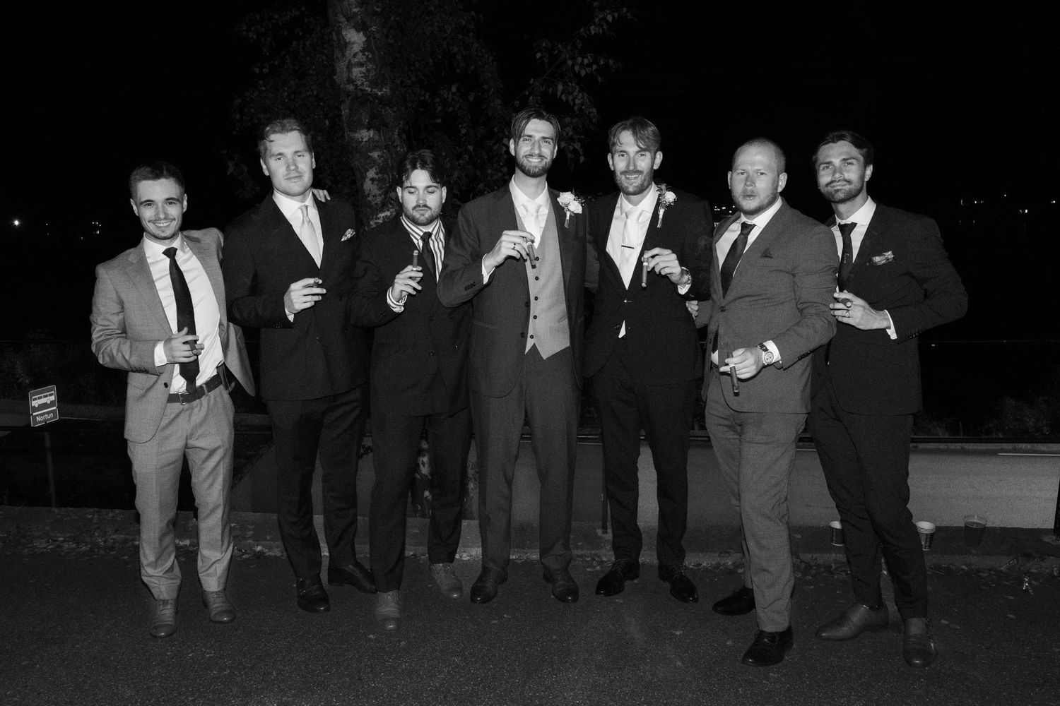 Black and white group photo of seven groomsmen in suits posing together at an outdoor evening wedding.
