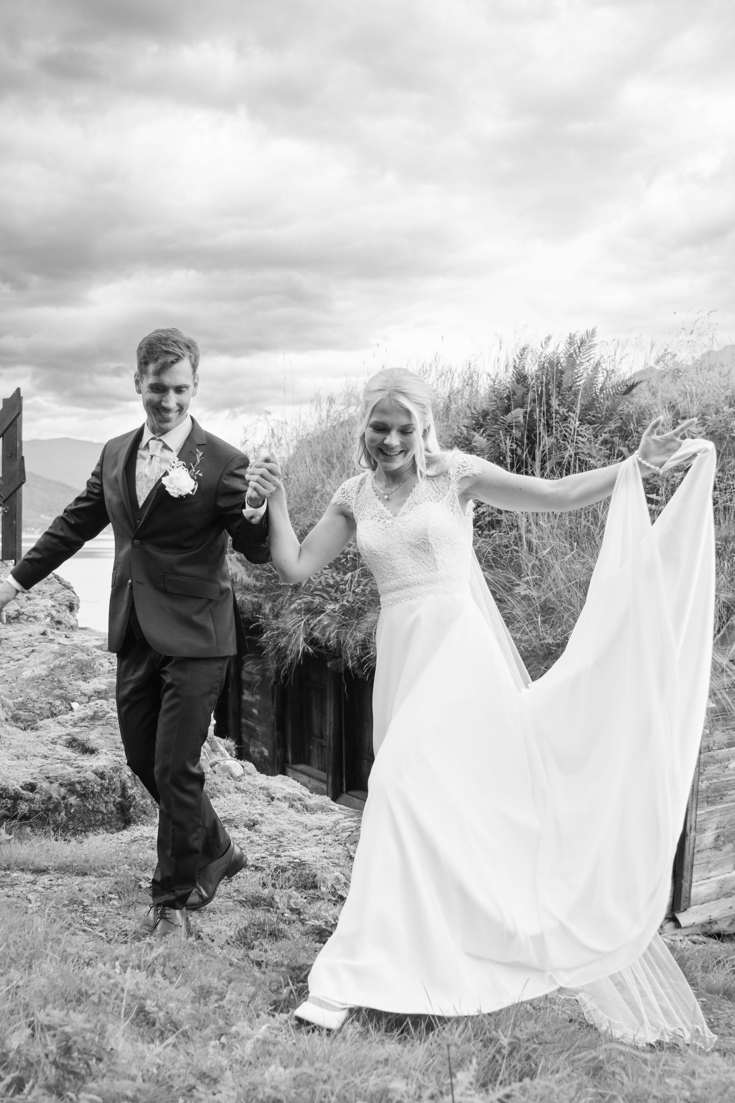 Black and white photo of smiling bride and groom walking hand in hand near a rustic hillside structure.