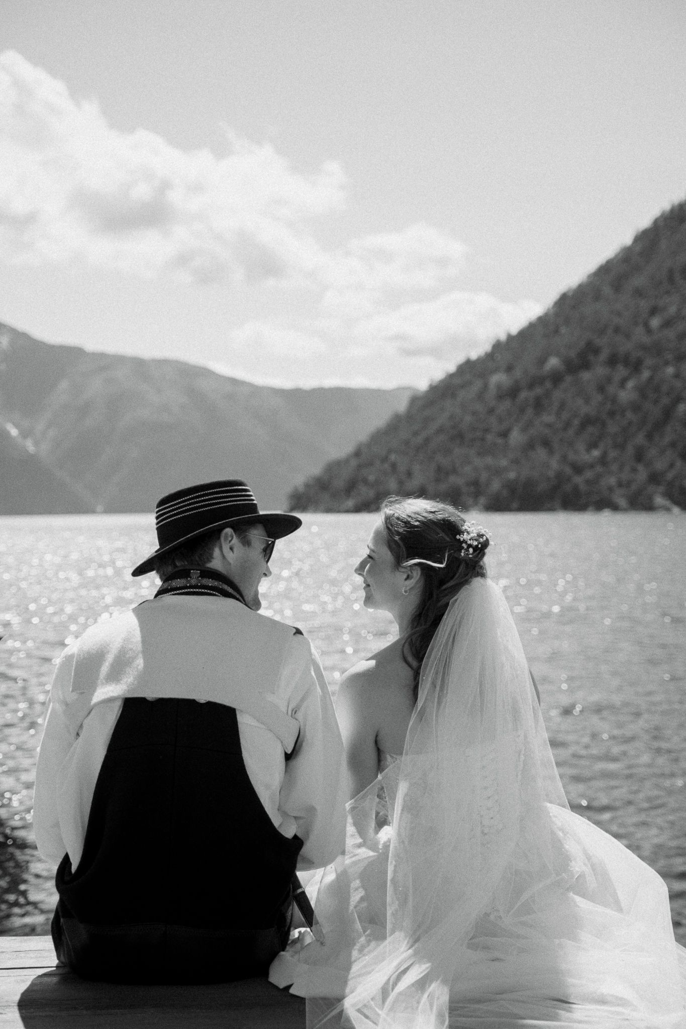 Bride and groom in traditional Norwegian bunad sit together on a dock gazing at a mountain fjord.
