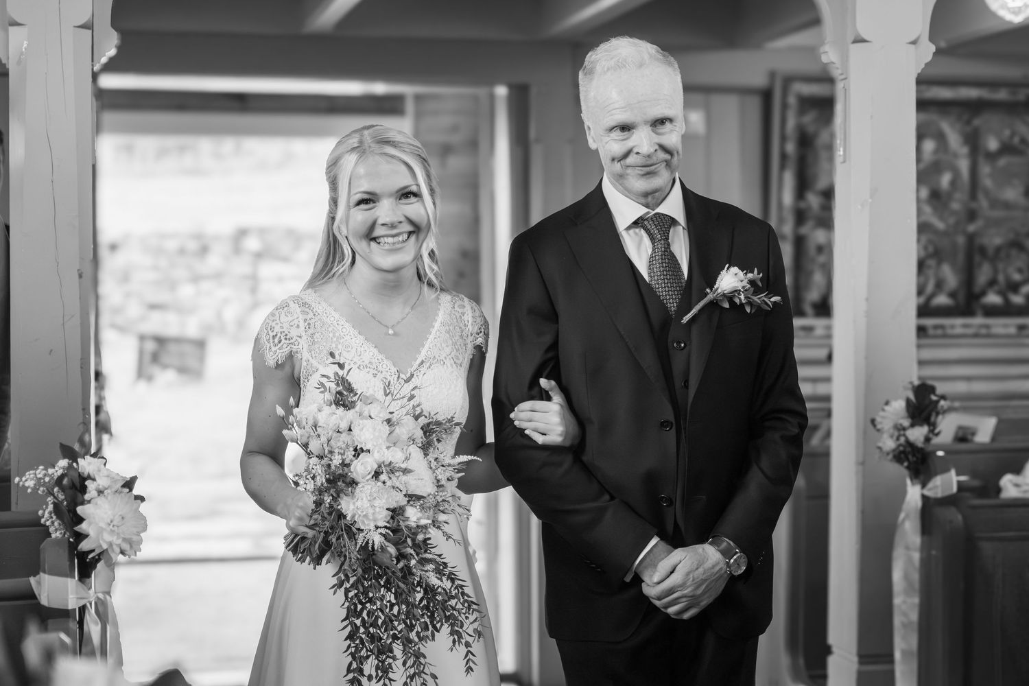 Bride walking down aisle with escort, smiling, holding floral bouquet in church.