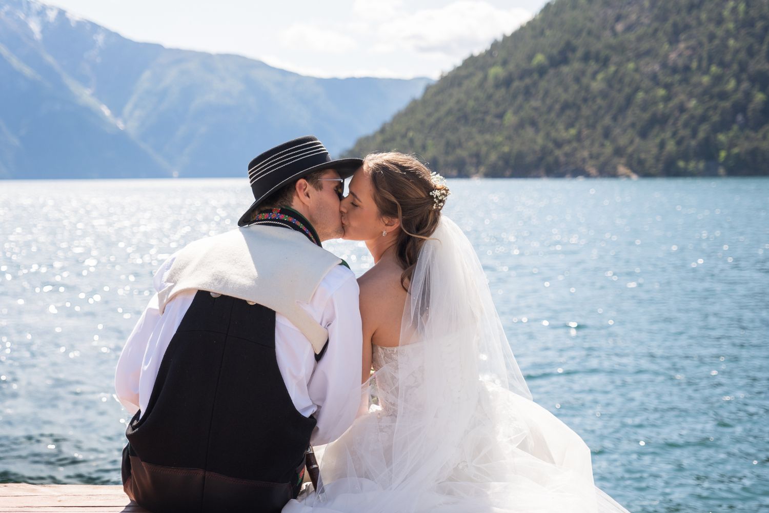 Bride and groom share a romantic kiss while seated on a dock with a sparkling mountain lake behind them.