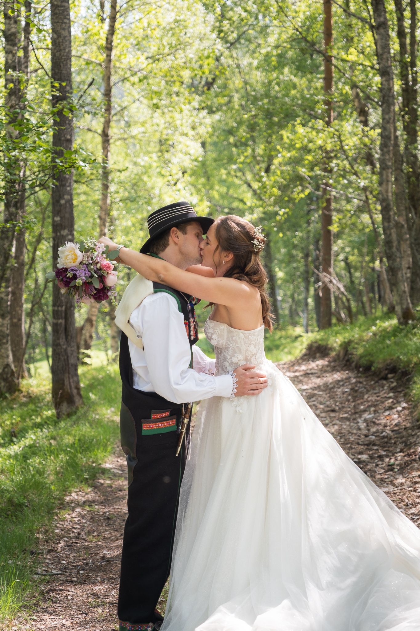 Bride and groom share a kiss on a sunlit forest path, groom holding a floral bouquet in traditional Norwegian bunad.