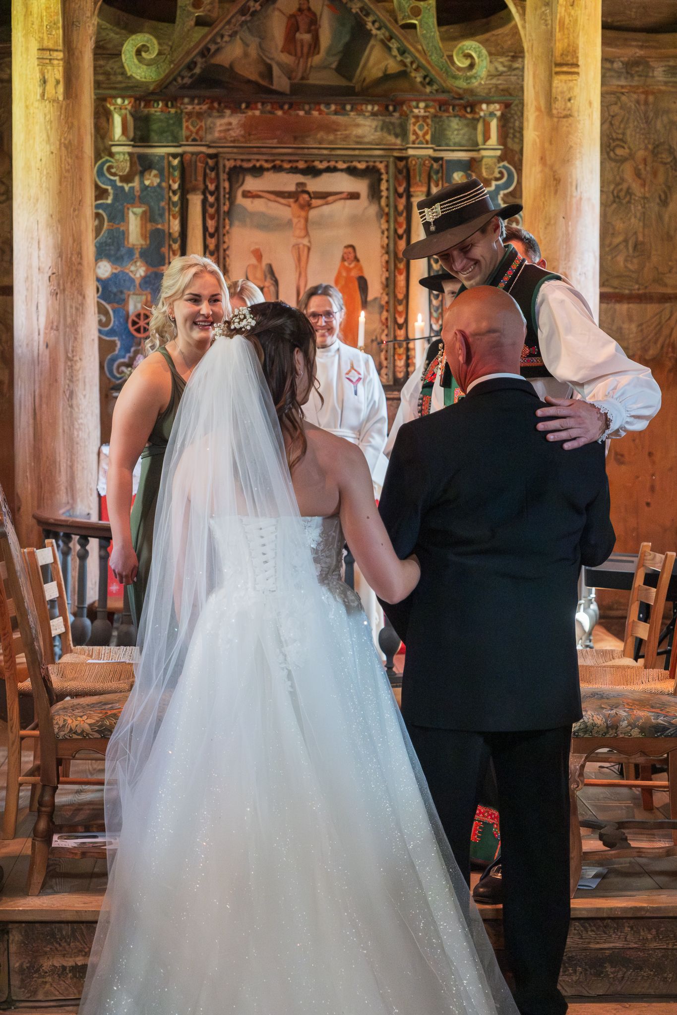 Bride arrives at altar greeted by groom in traditional hat inside a beautifully decorated historic wooden church.