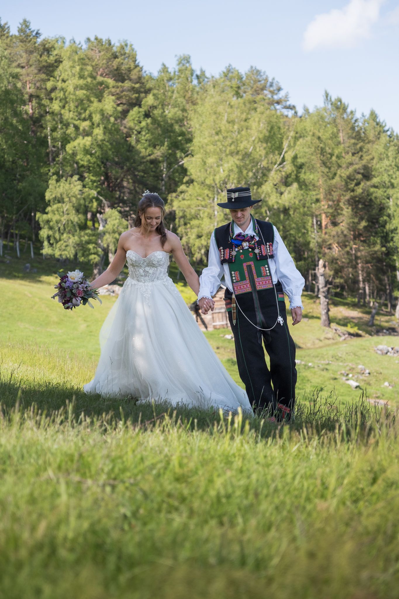 Bride in white gown and groom in traditional Norwegian bunad walk hand in hand through a green meadow.