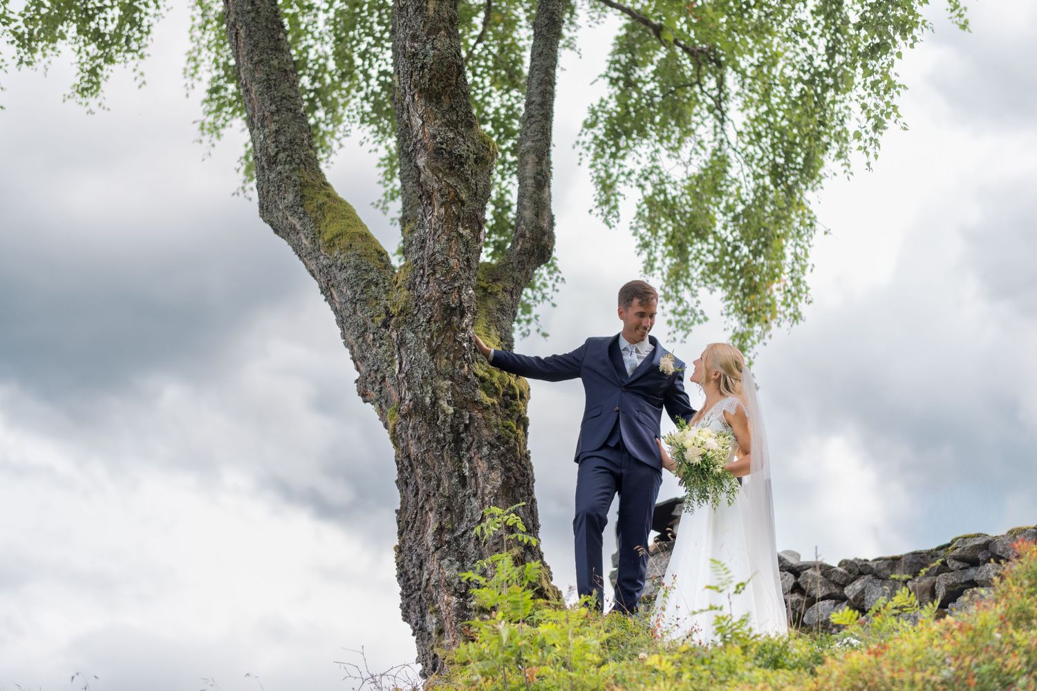 Bride and groom stand together under large birch tree on hillside with dramatic cloudy sky backdrop.
