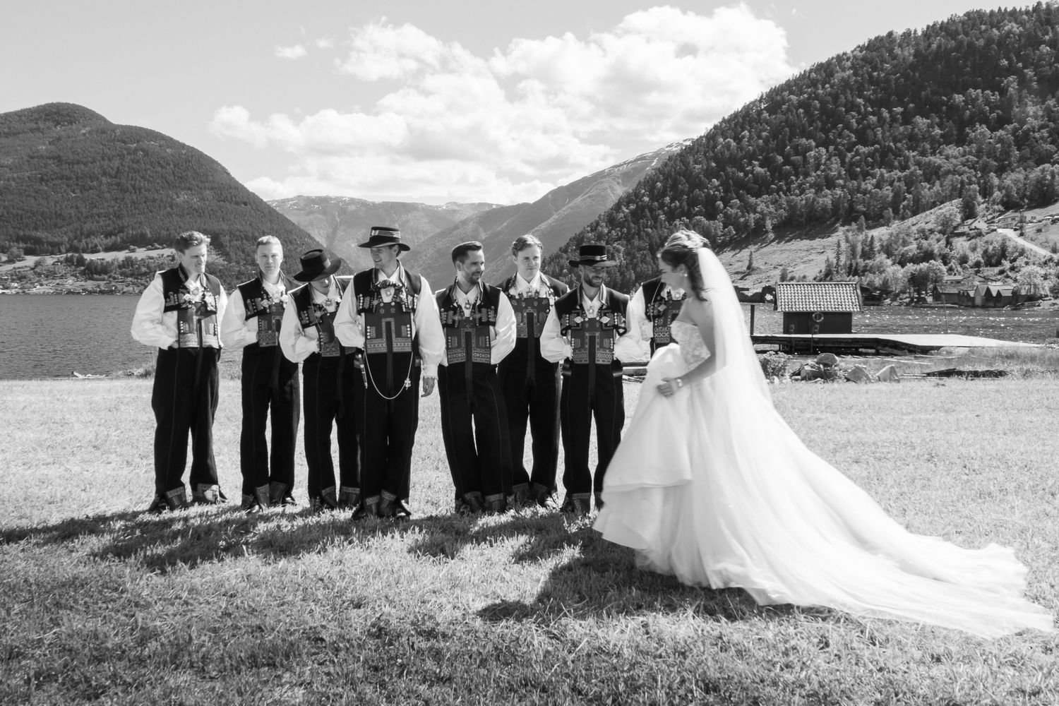 Groomsmen in Norwegian bunad attire holding bouquets beside a bride in a white gown by a mountain lake.
