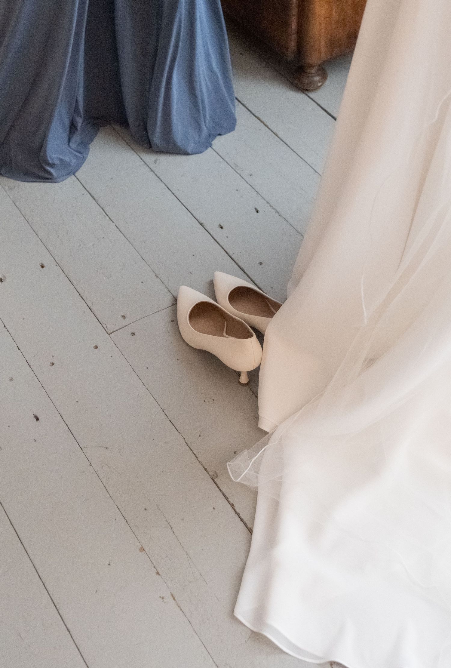 Nude bridal heels placed on rustic white wooden floor beside flowing white wedding dress and blue bridesmaid gown.