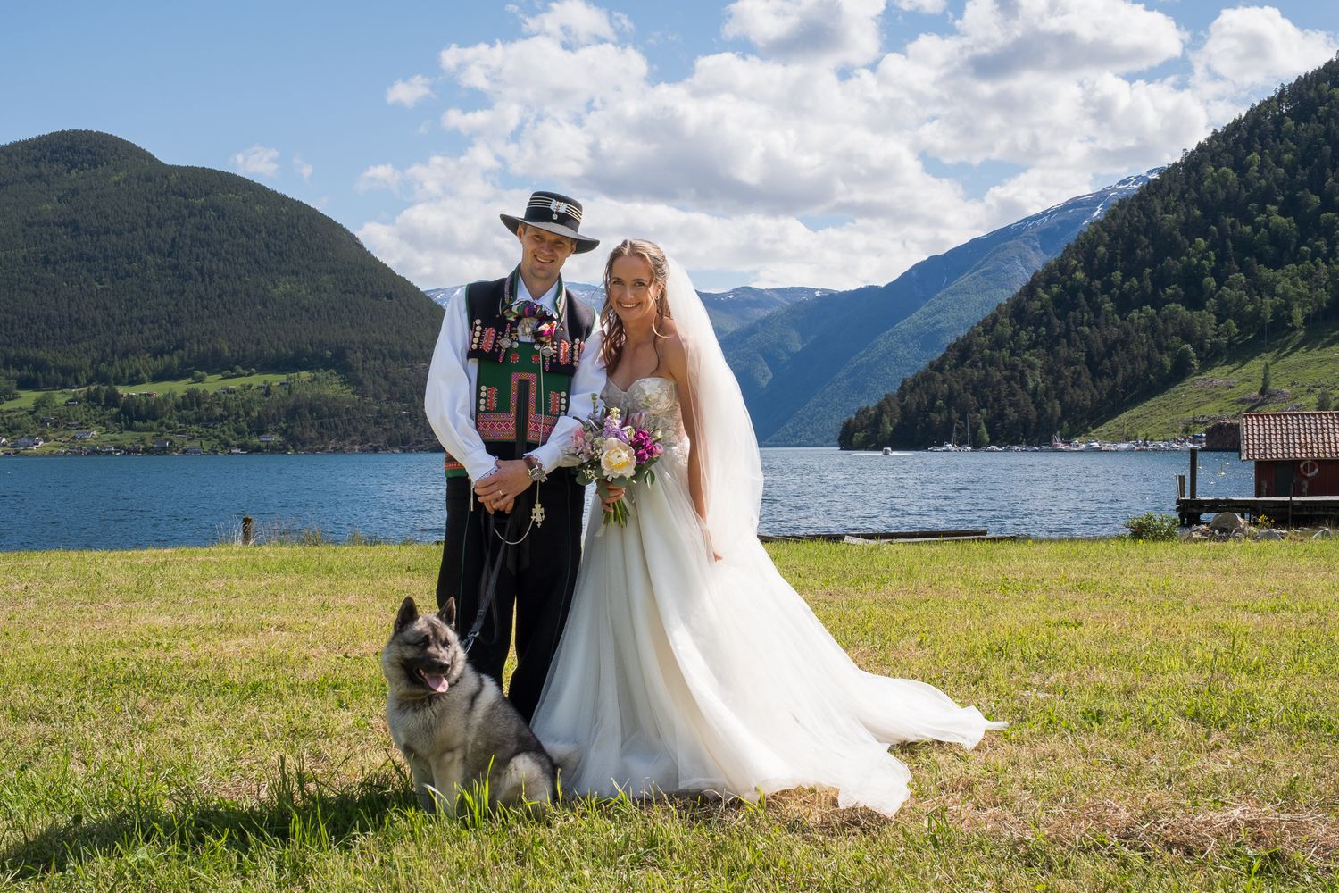 Norwegian groom in traditional bunad and bride in white gown pose with a dog by a mountain fjord.