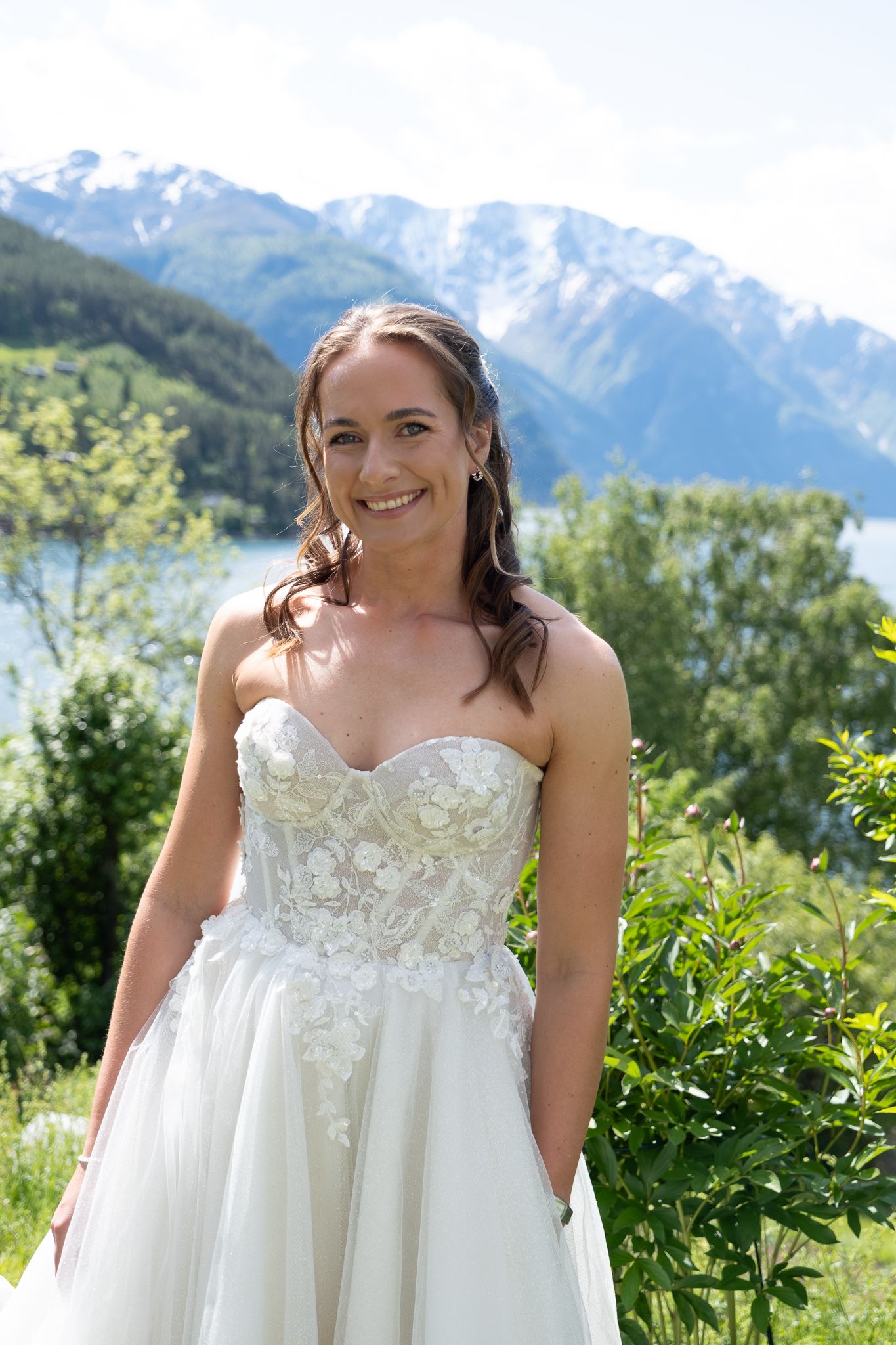 Smiling bride in strapless lace wedding gown poses outdoors with alpine lake and snow-capped mountains behind her.