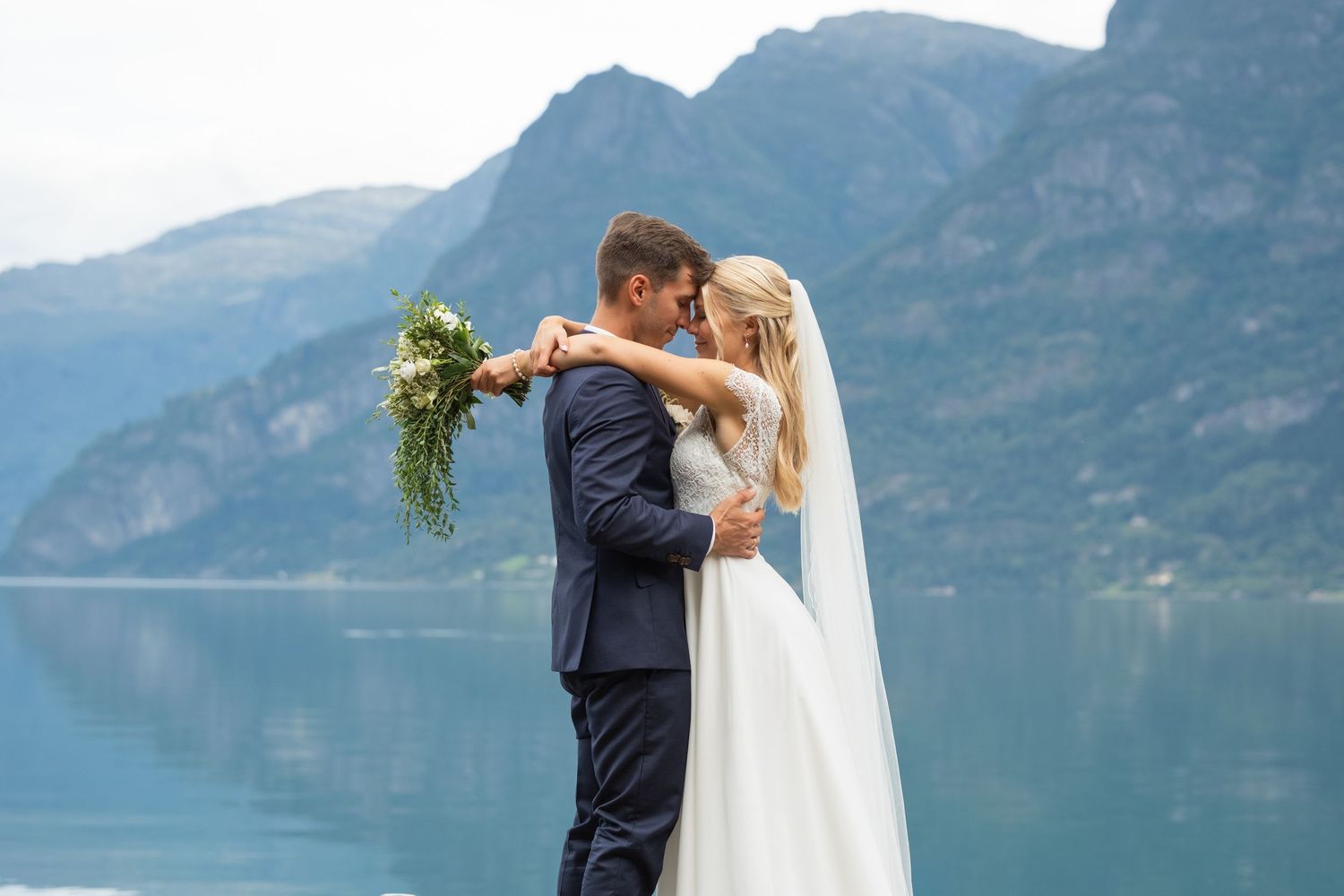 A bride and groom share a tender embrace by a serene fjord with majestic mountains behind them.