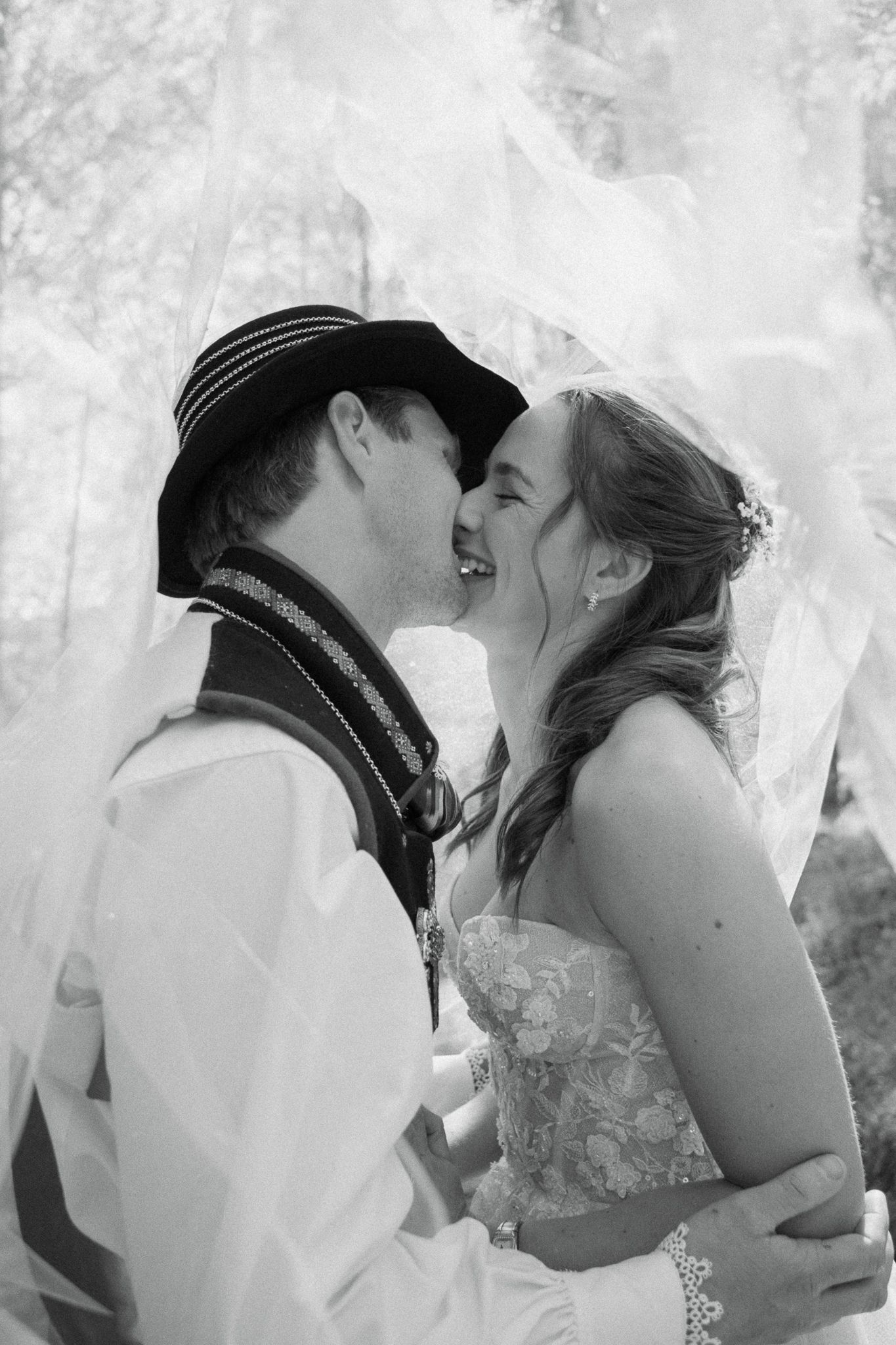 Black and white photo of a bride and groom sharing a joyful kiss under a flowing veil outdoors.