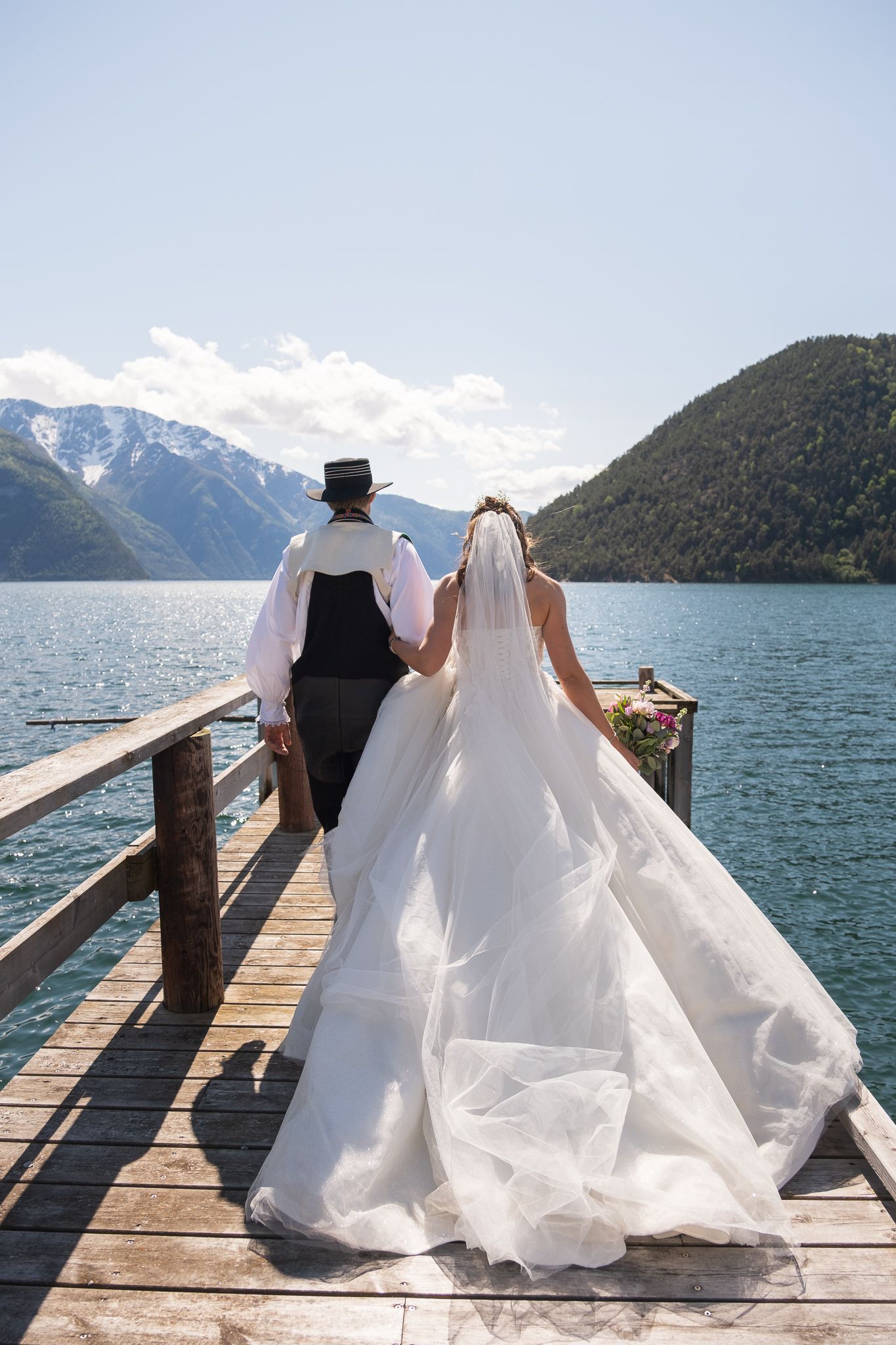 Bride and groom stand on a wooden dock overlooking a serene mountain lake on their wedding day.