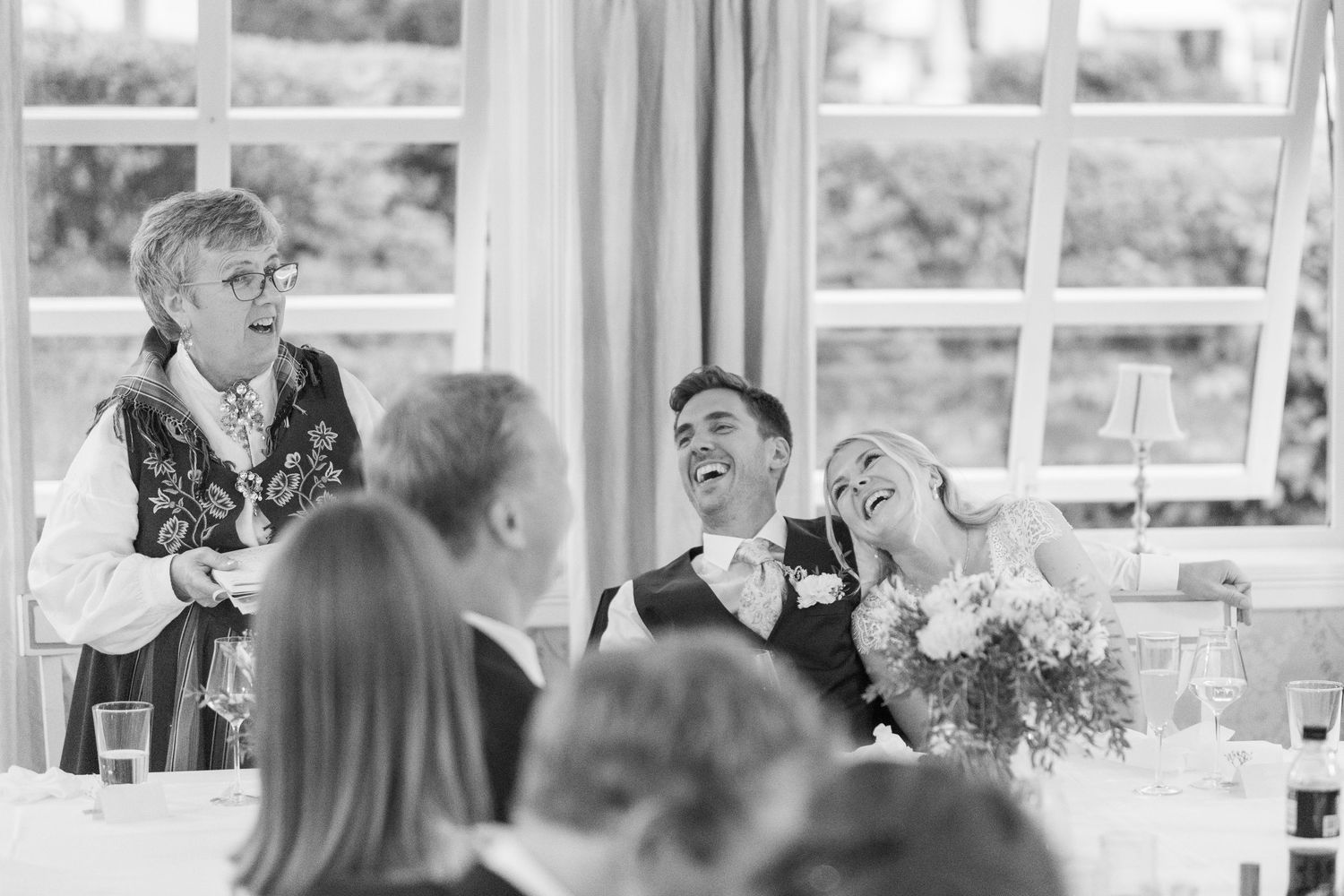Black and white candid of bride and groom laughing heartily during a speech at their wedding reception.