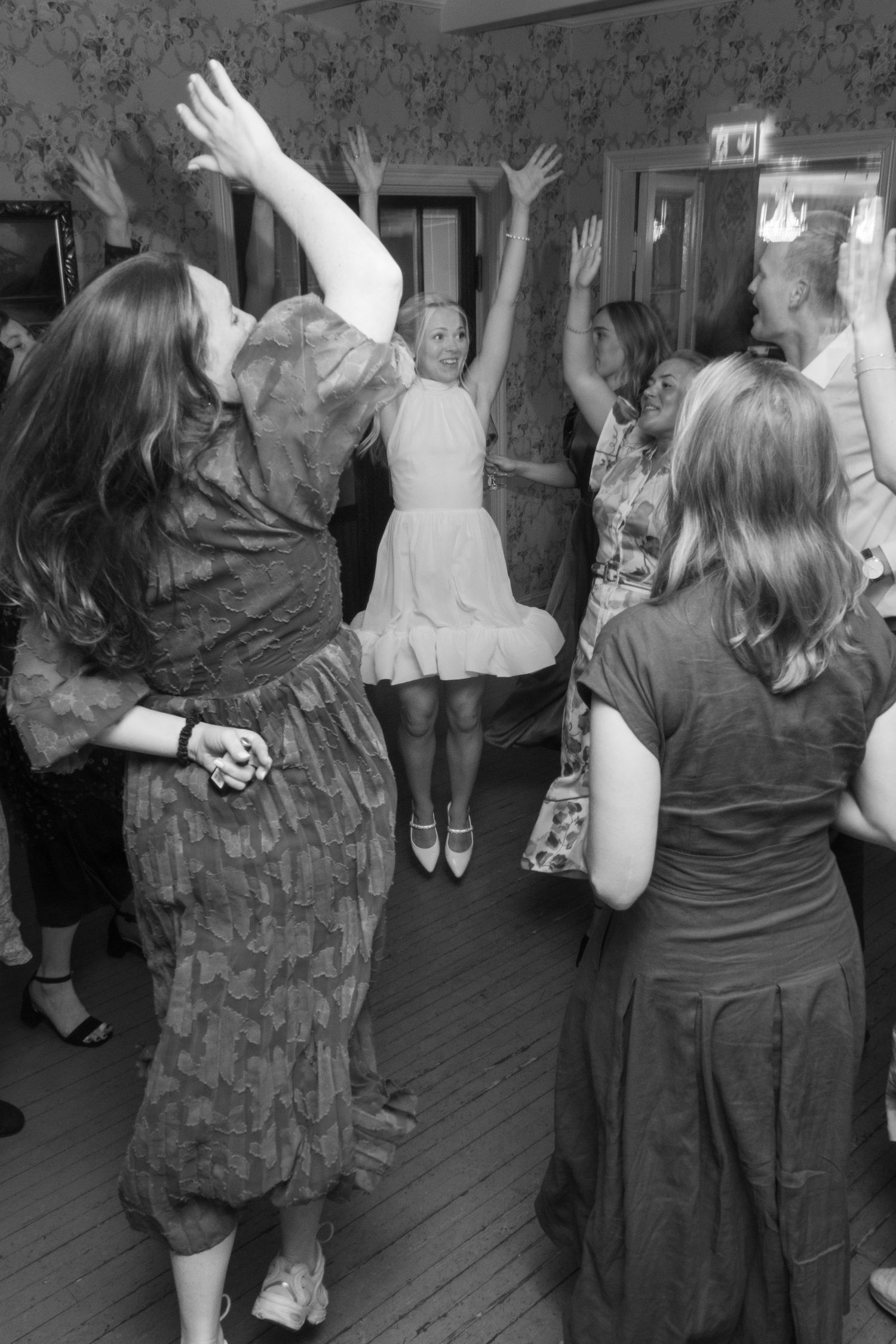 Bride in white dress jumping with arms raised surrounded by dancing guests at wedding reception.