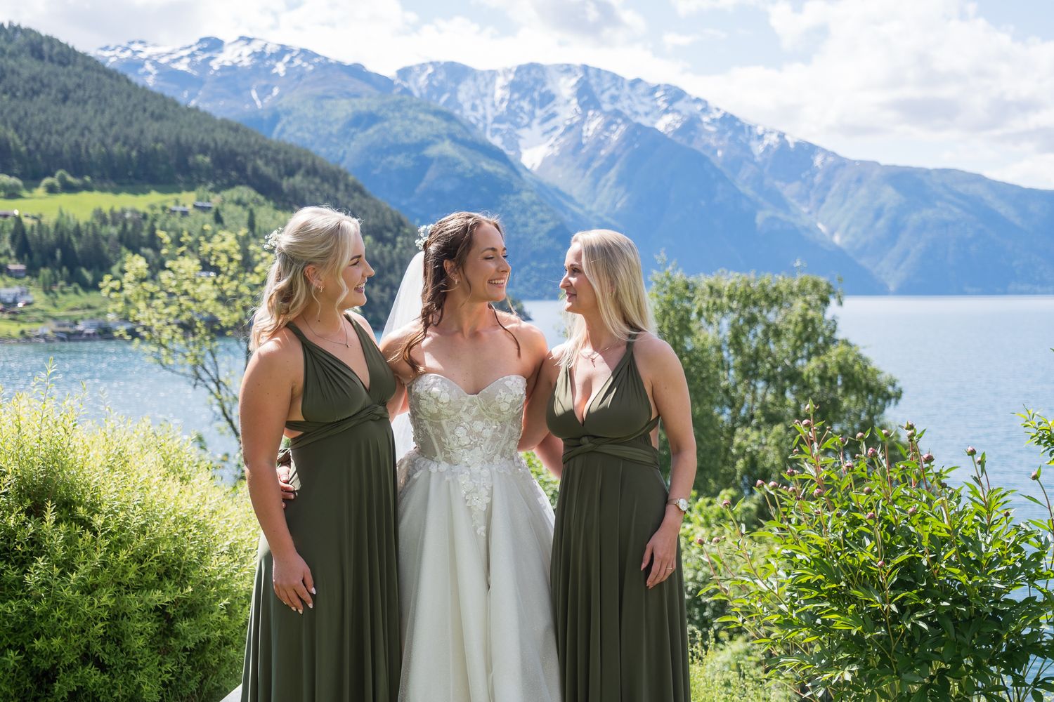 Bride in white gown with two bridesmaids in olive green dresses by a mountain lake in Norway.