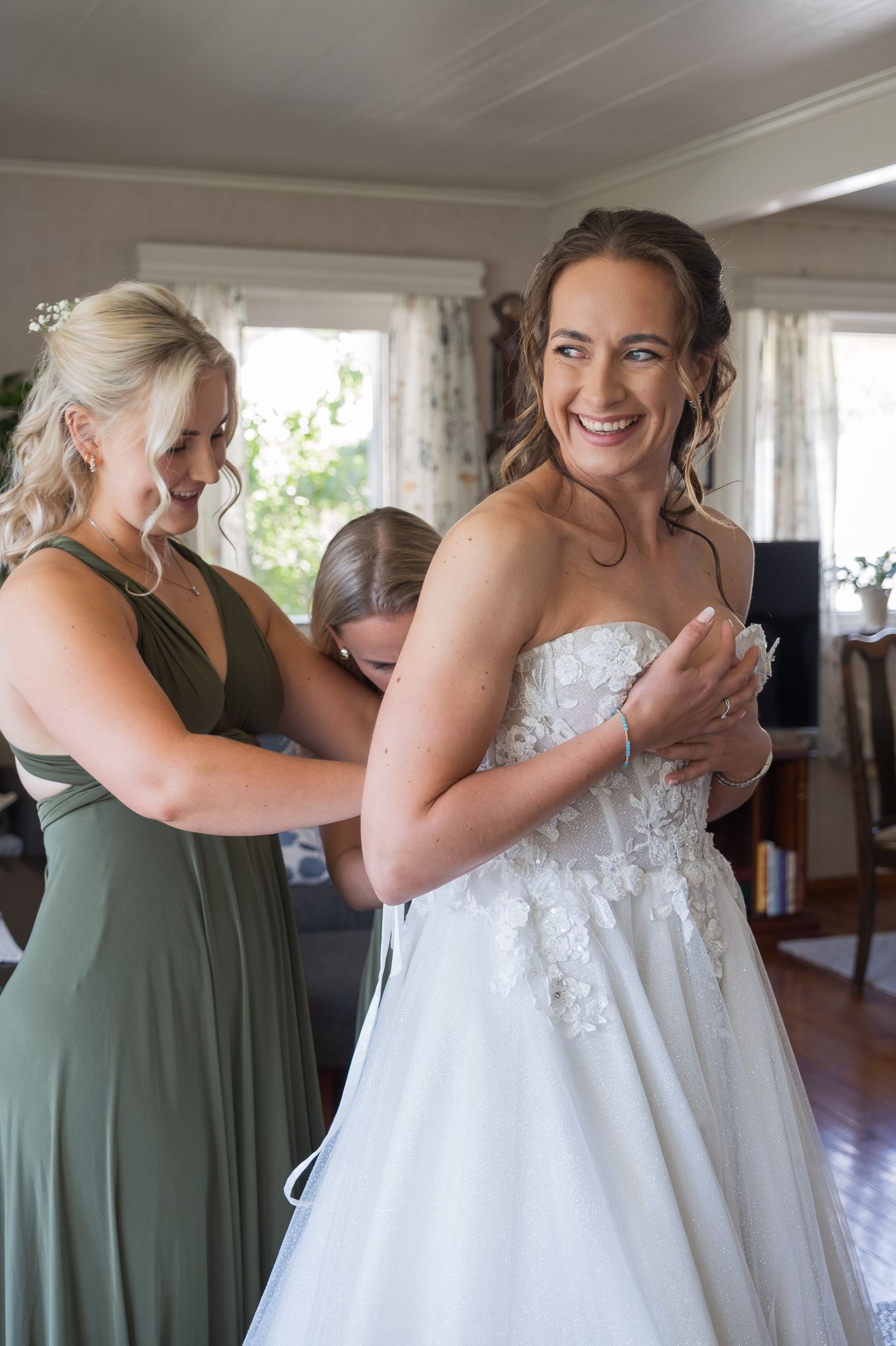 Smiling bride in strapless lace wedding dress being laced up by bridesmaids in sage green gowns.