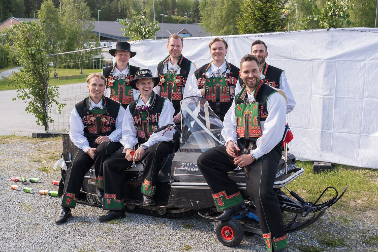 Group of men in matching traditional Norwegian bunad vests and hats pose together on a snowmobile outdoors.