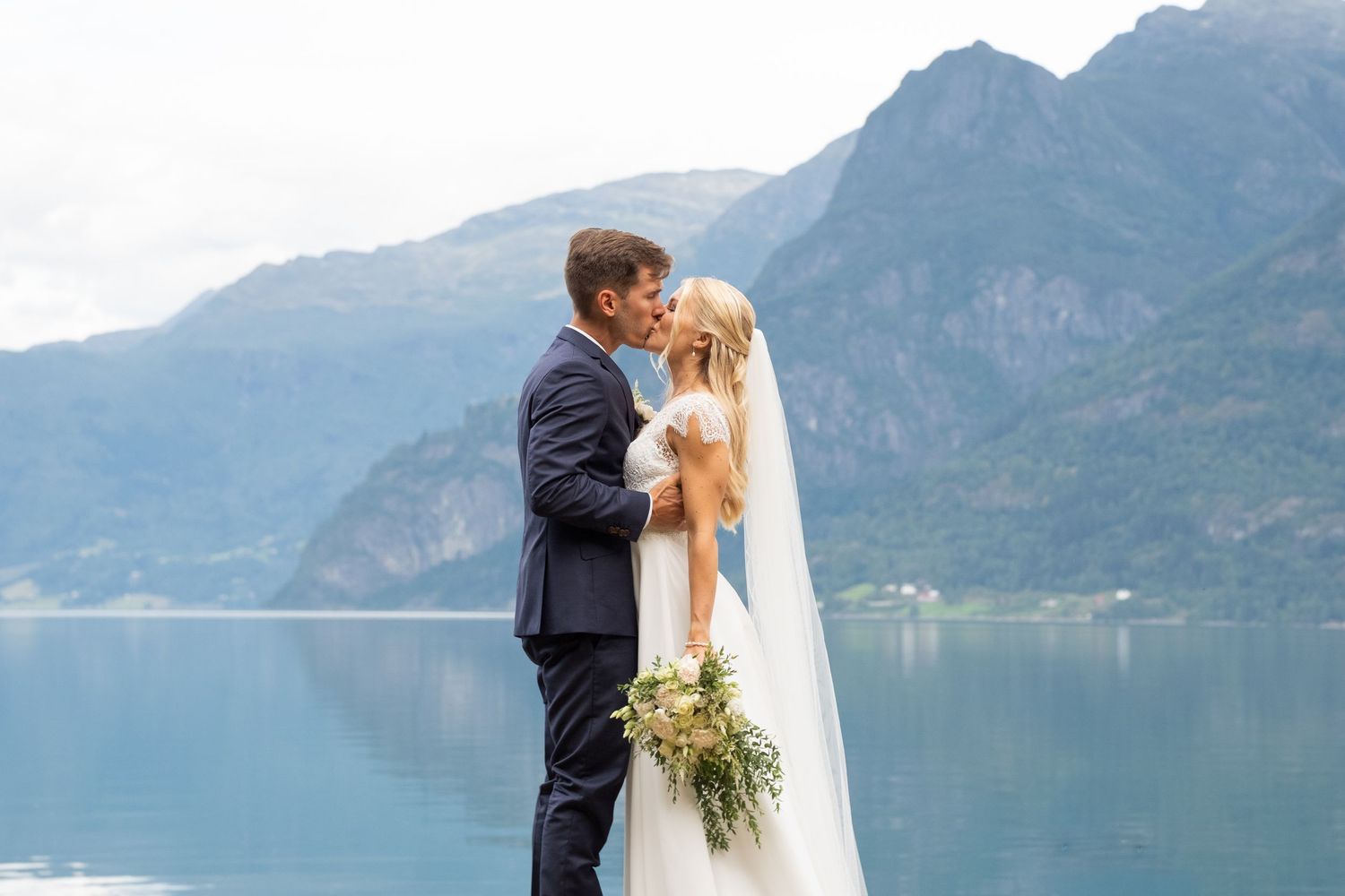 Bride and groom share a romantic kiss by a scenic fjord with mountain backdrop on their wedding day.