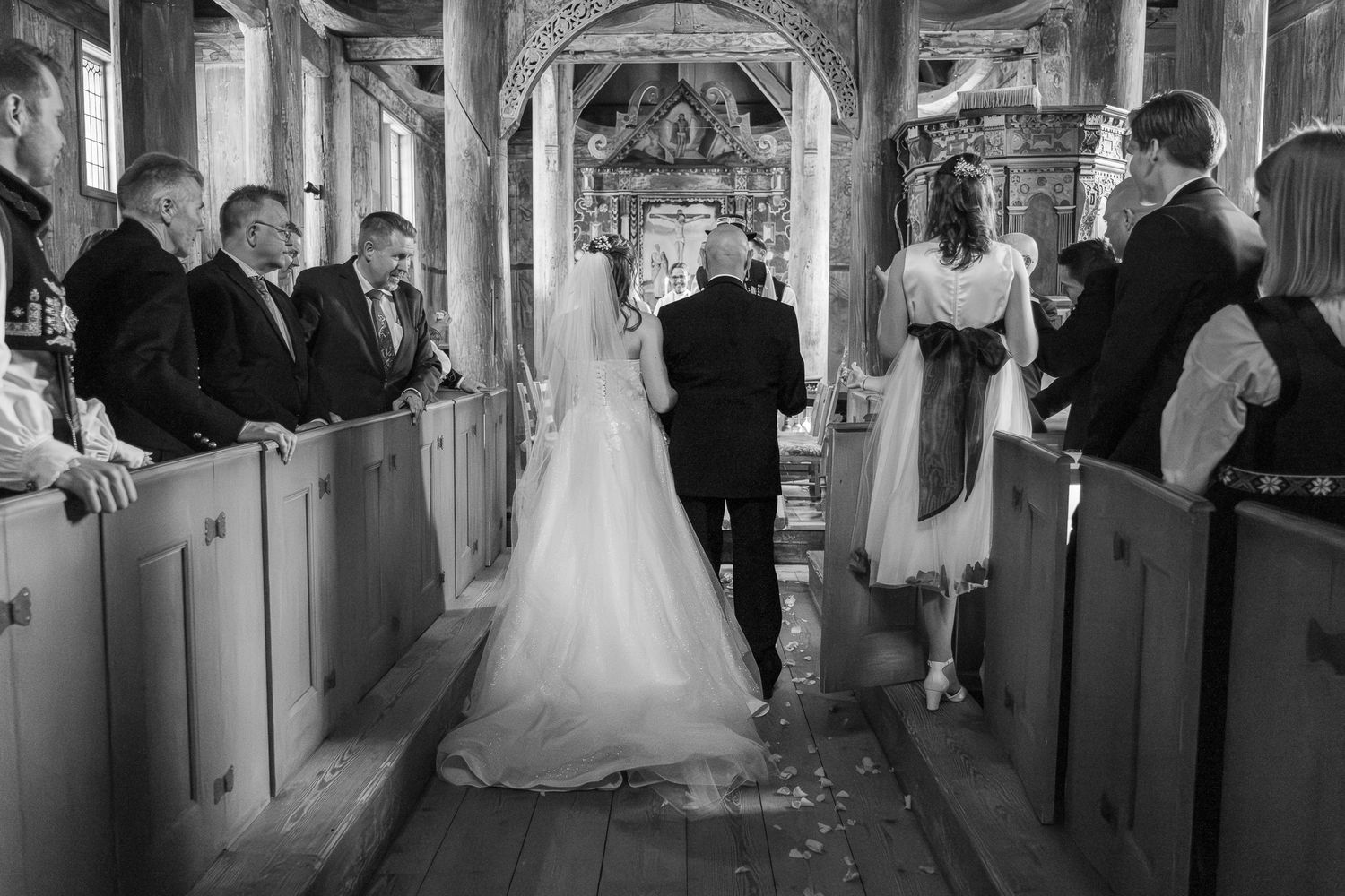 Black and white image of bride walking down the aisle of a rustic wooden church with guests standing in pews.