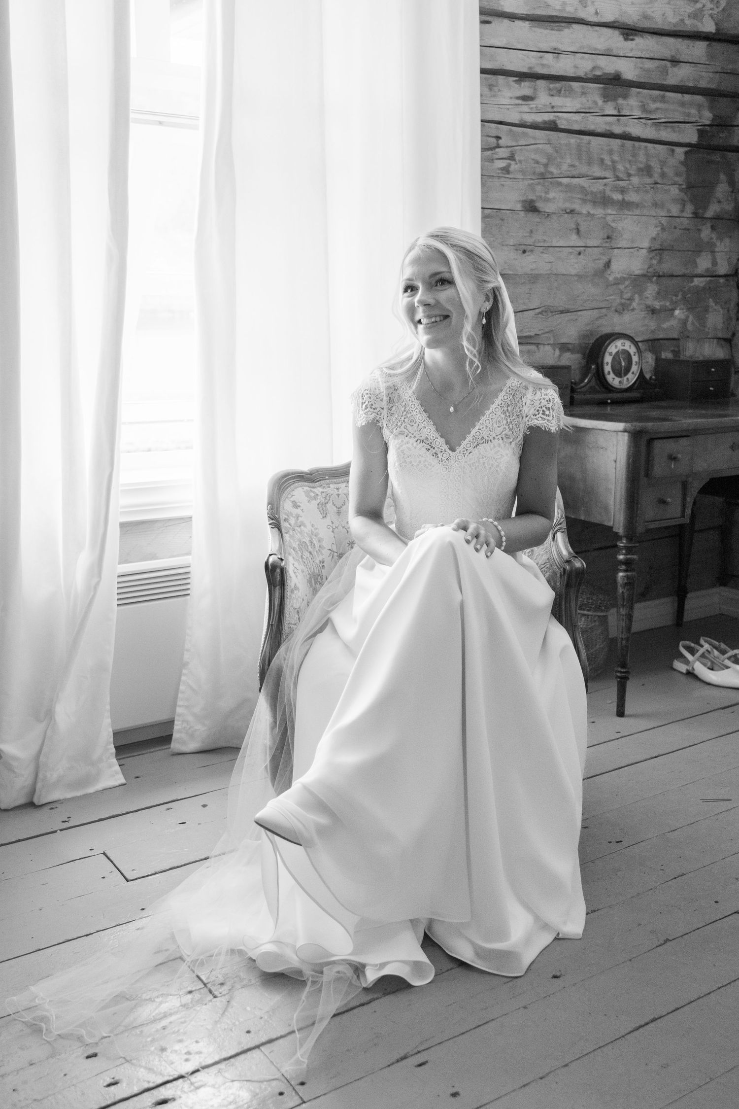 Elegant bride in lace wedding gown sitting in vintage chair smiling by window in black and white photo.