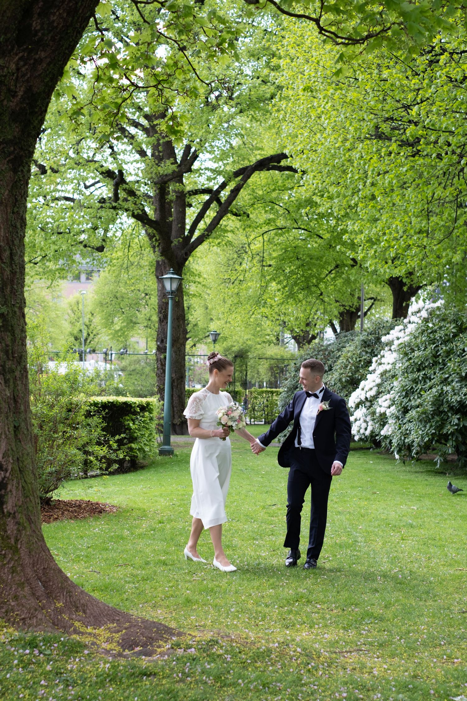 Bride and groom walking hand in hand through lush green park beneath large leafy trees in spring.