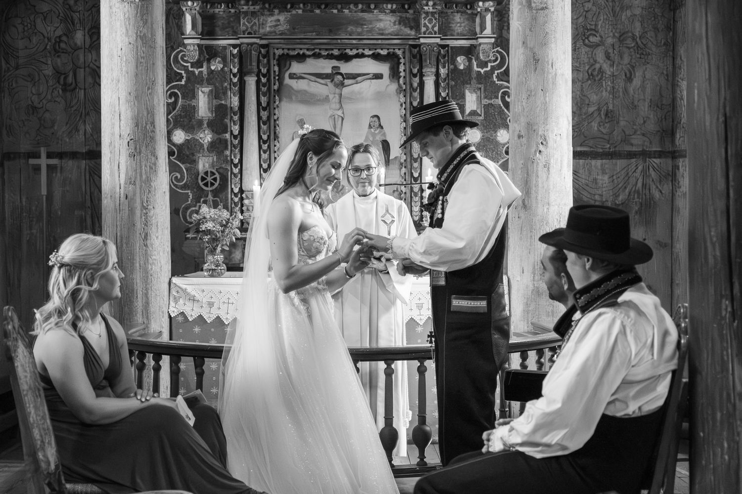 Black and white photo of bride and groom exchanging vows at the altar of a historic wooden church with witnesses.