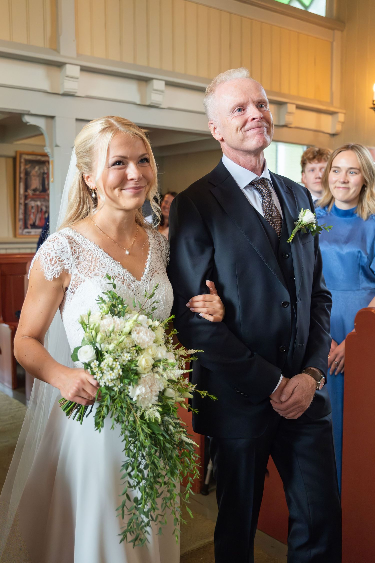 Smiling bride in lace wedding dress walks down the aisle holding bouquet beside suited man in church.