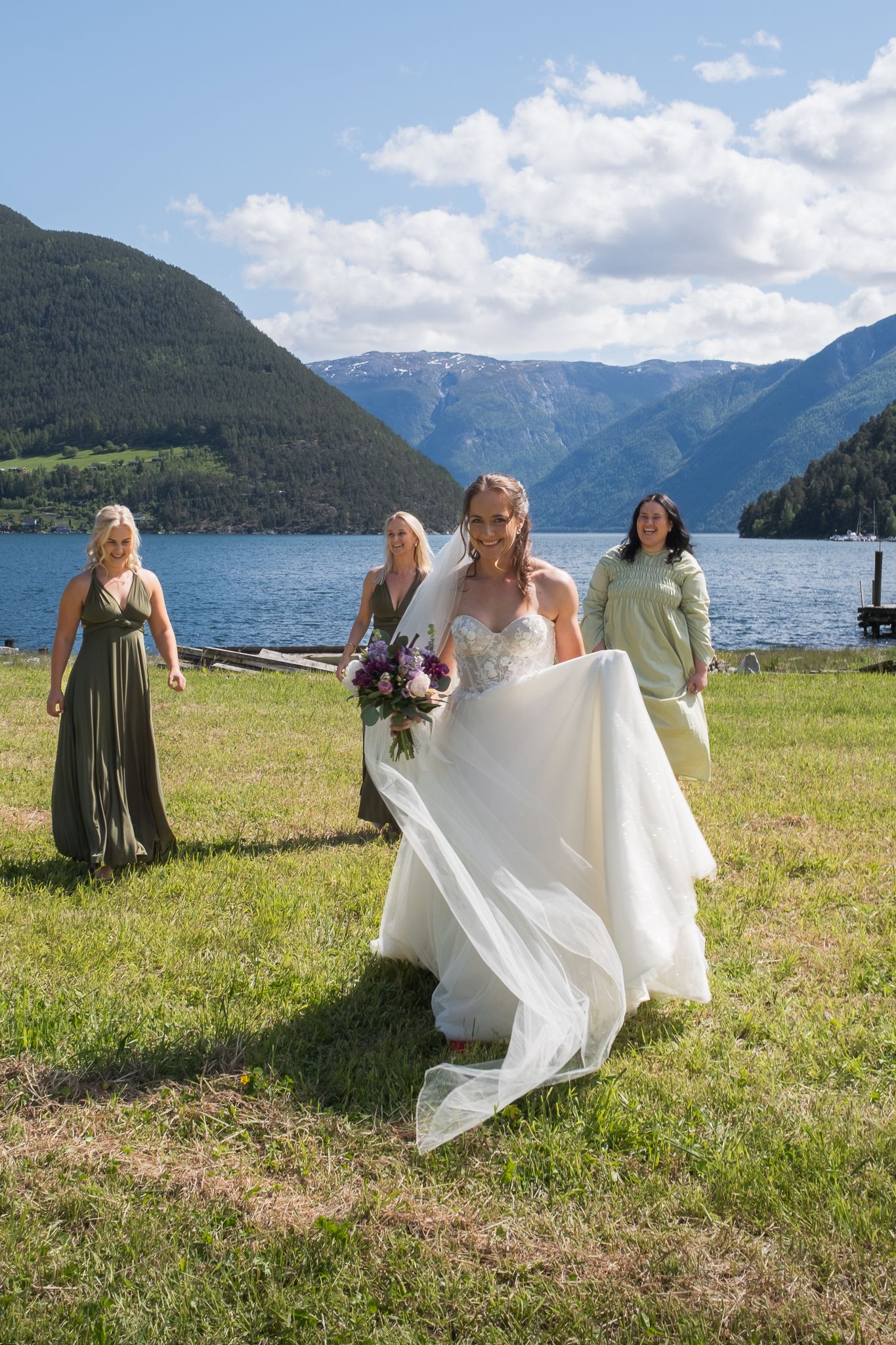 Smiling bride in white gown carries her dress while bridesmaids follow along a scenic mountain lakeside.