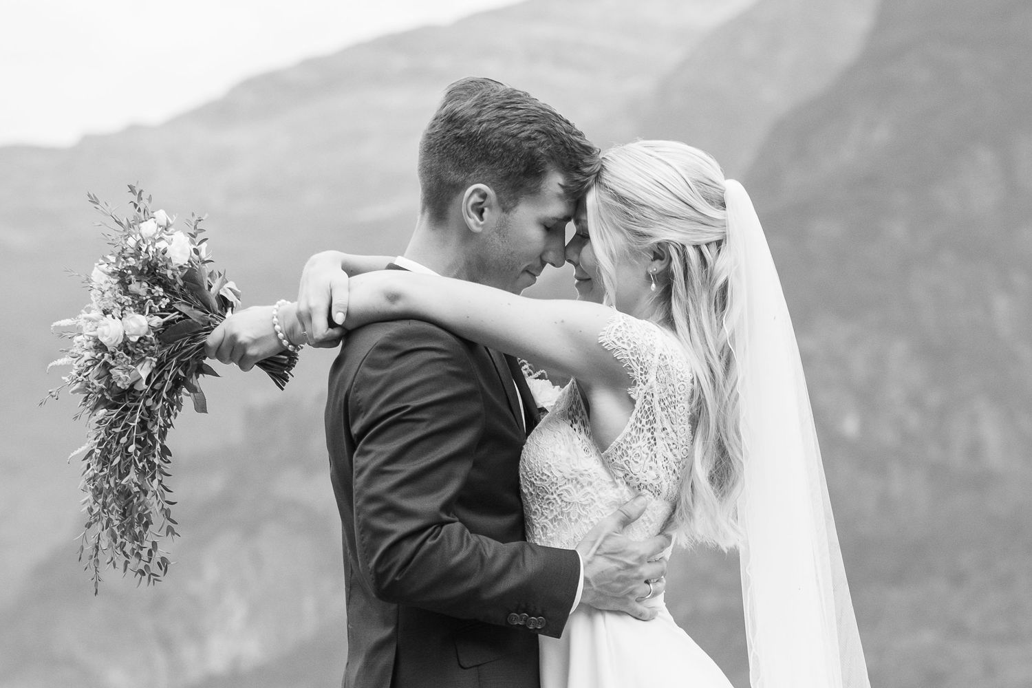 Black and white wedding portrait of couple touching foreheads tenderly with mountain scenery behind them.
