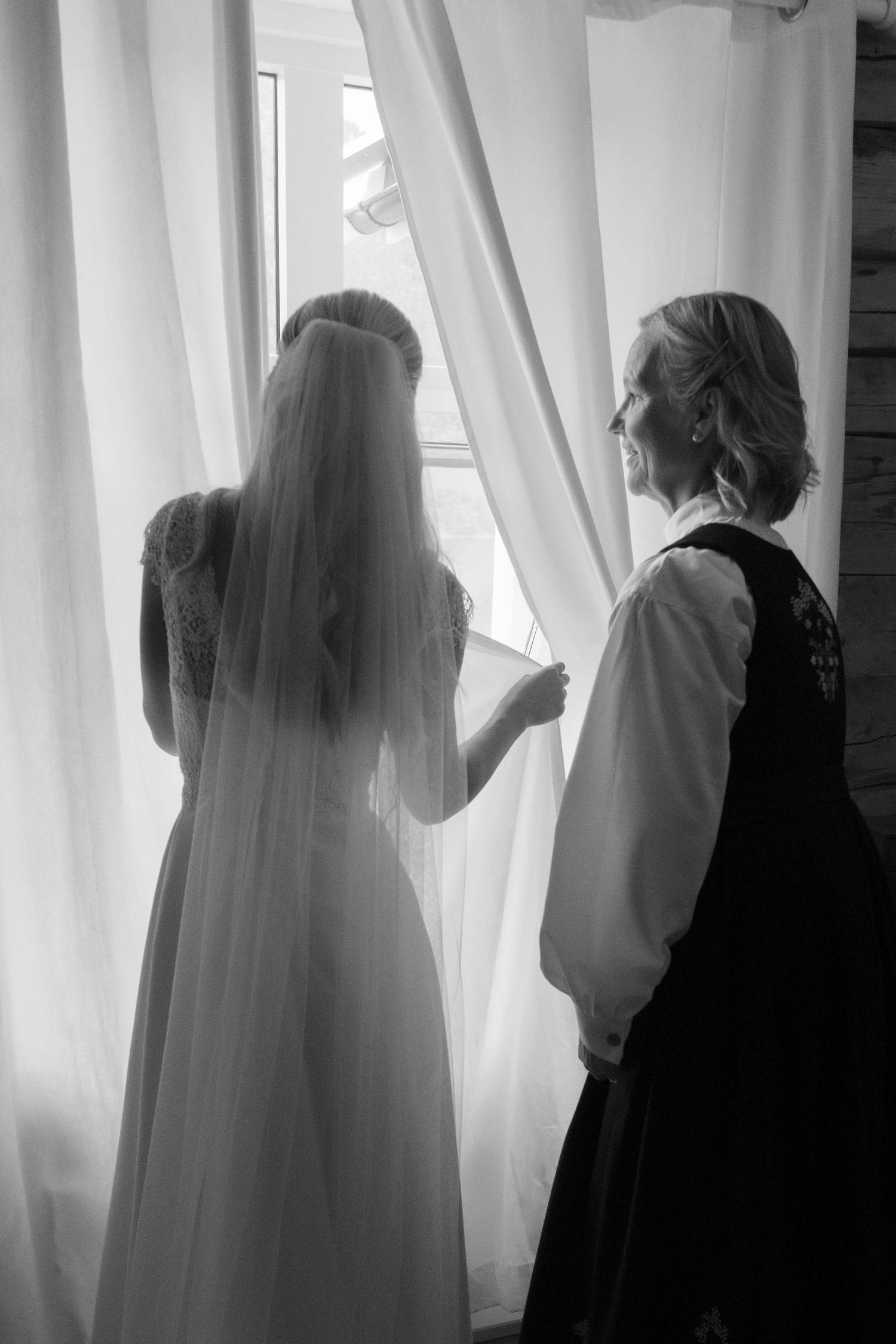 Bride in wedding gown and veil standing by a window with a woman in traditional dress in black and white.
