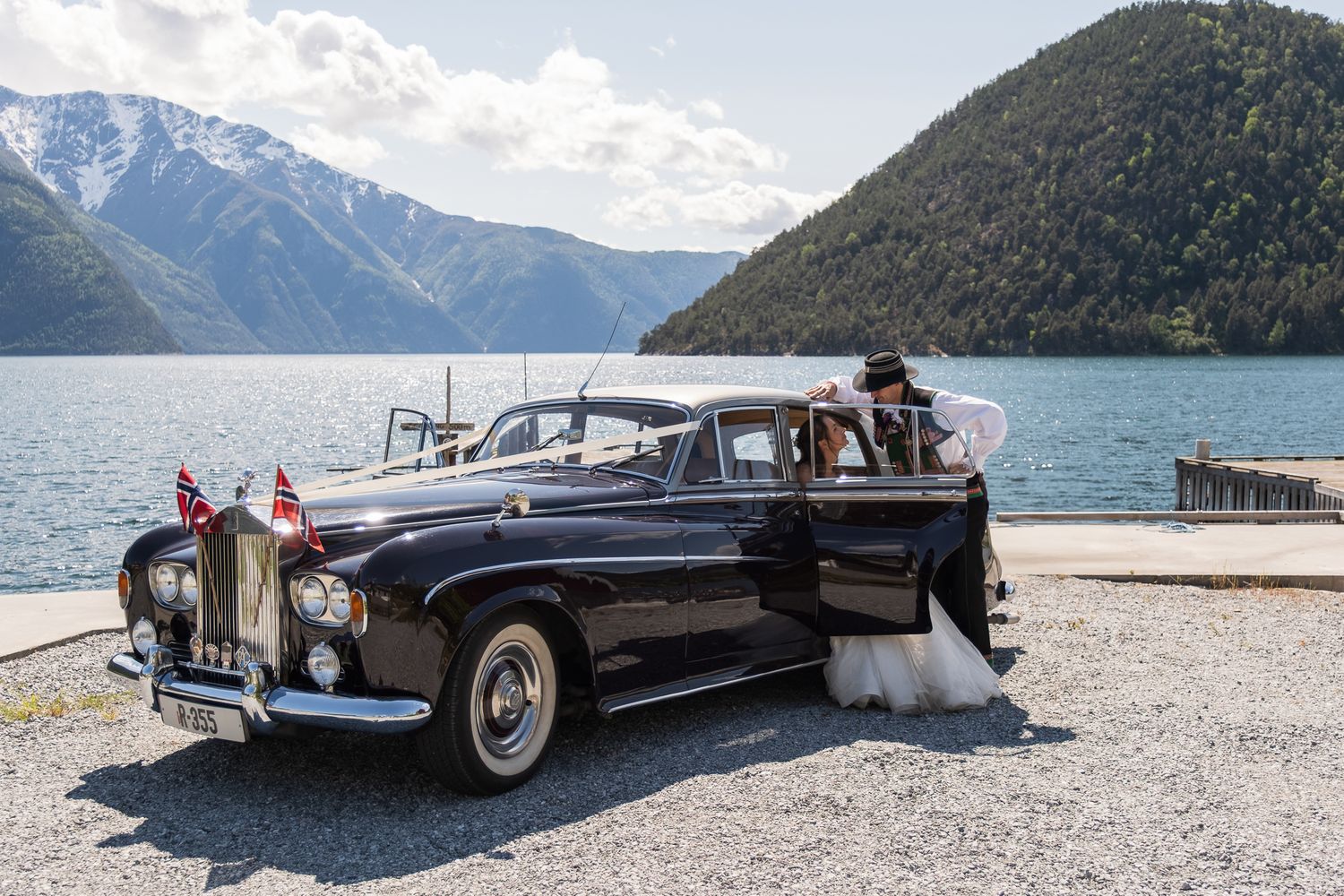 Bride and groom pose beside a classic vintage Rolls-Royce decorated with flags near a mountain lake.