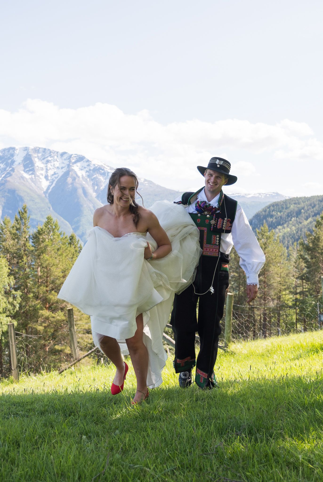 Bride and groom in Norwegian bunad walk across a sunny alpine meadow with snow-capped mountains in the background.