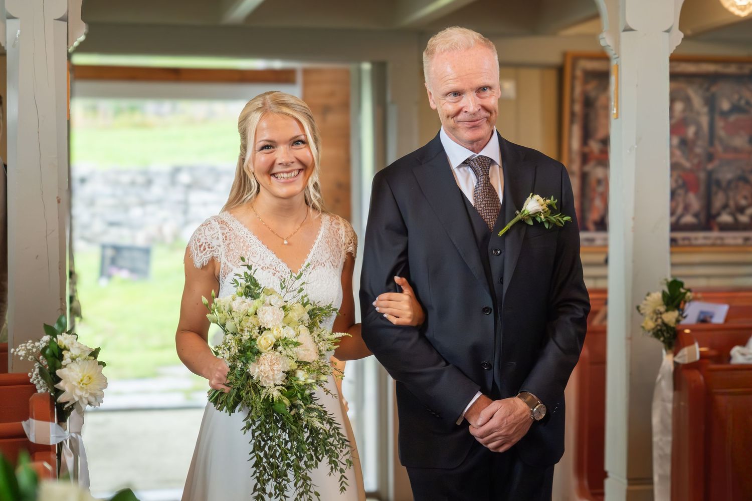 Smiling bride in lace gown holding bouquet walking down aisle with escort in navy suit.
