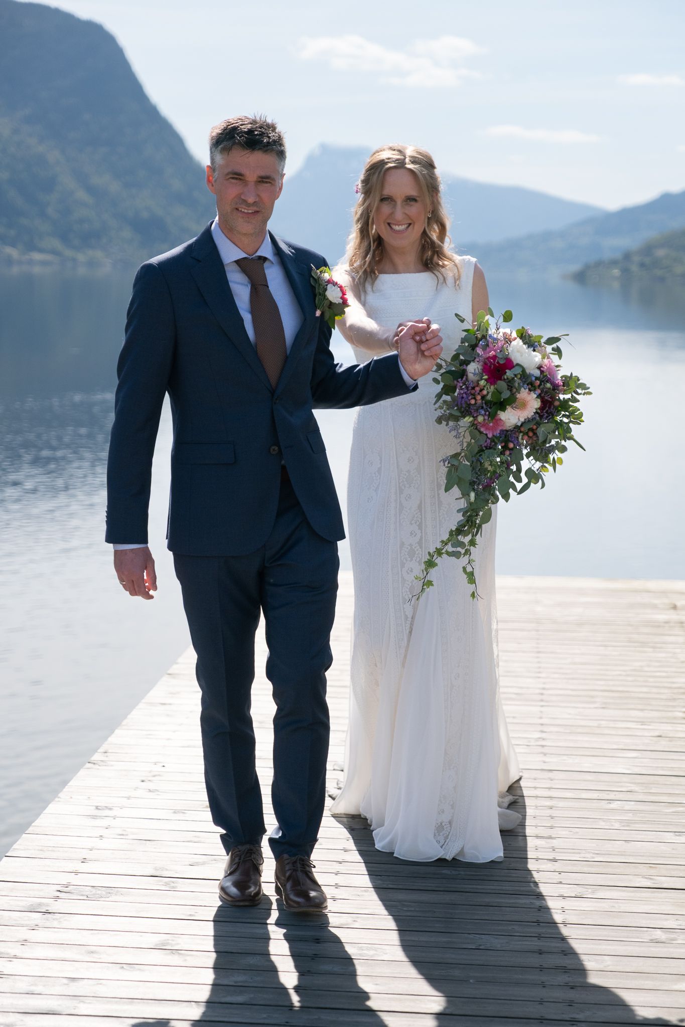 Bride and groom holding hands on a wooden dock with a calm fjord and mountains behind them.