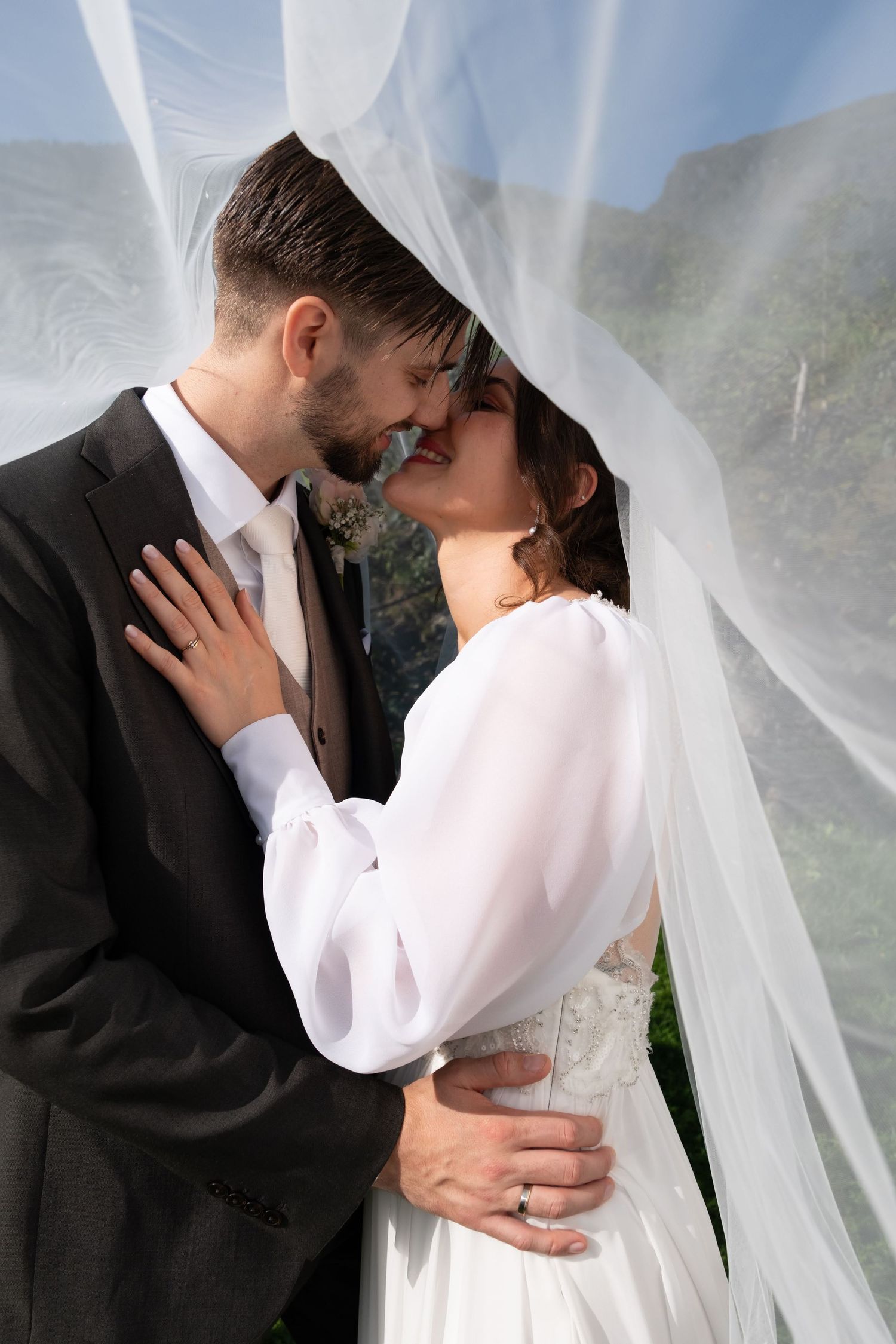 Bride and groom sharing a romantic moment under flowing wedding veil with mountain waterfall backdrop.