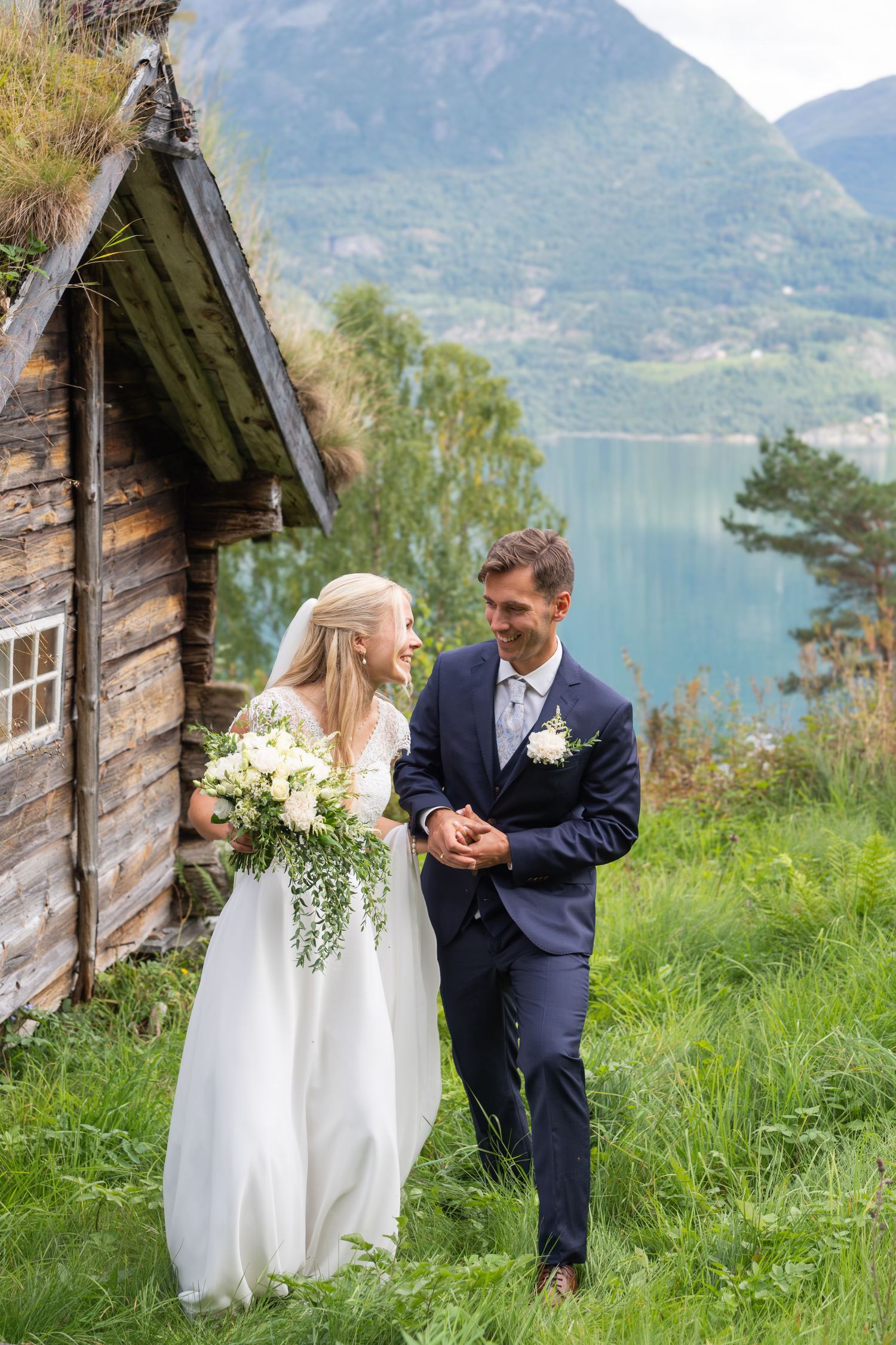 Smiling bride and groom walk together beside rustic log cabin with scenic Norwegian fjord behind.