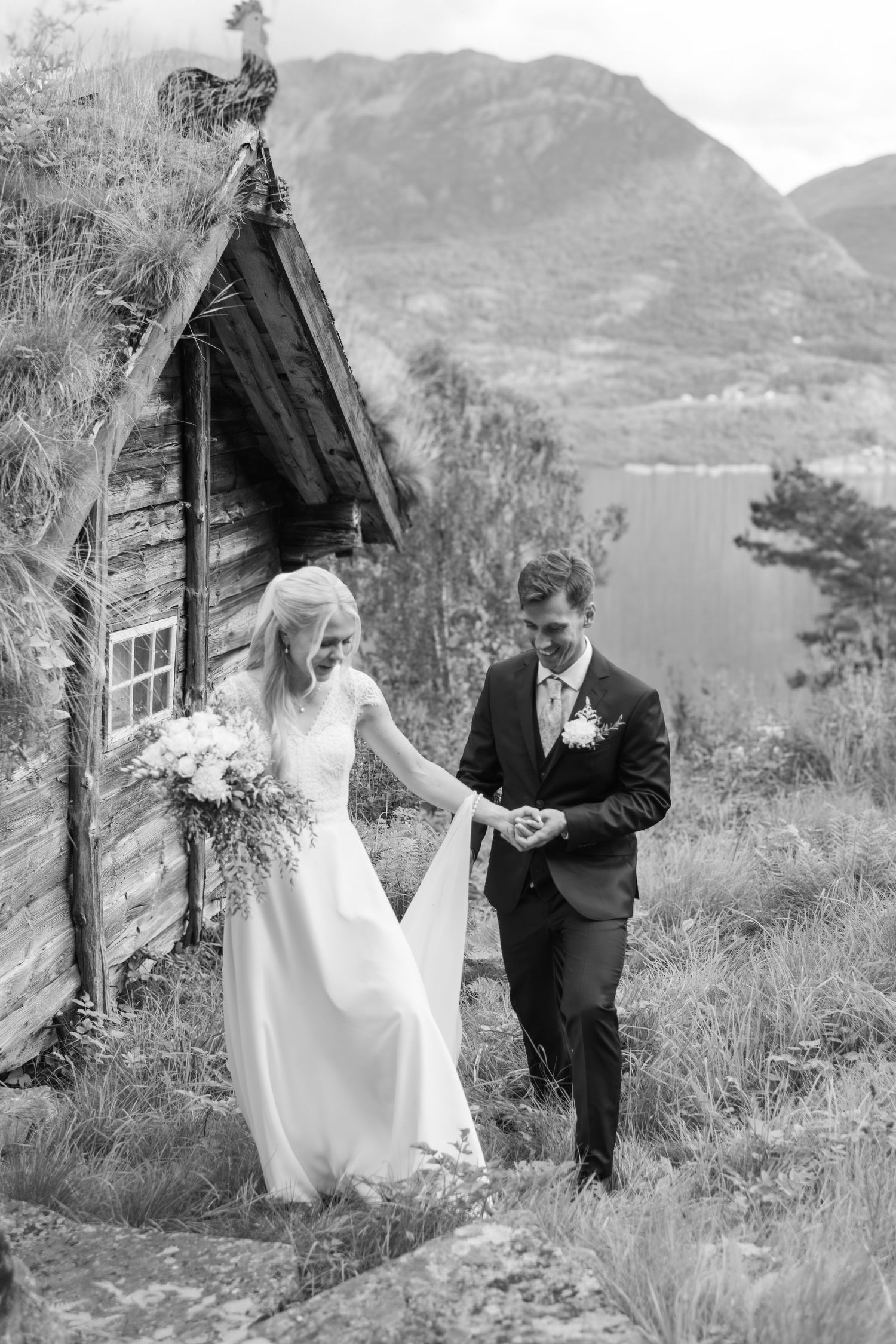 Bride and groom walk hand in hand beside rustic sod-roof cabin with mountain lake backdrop.