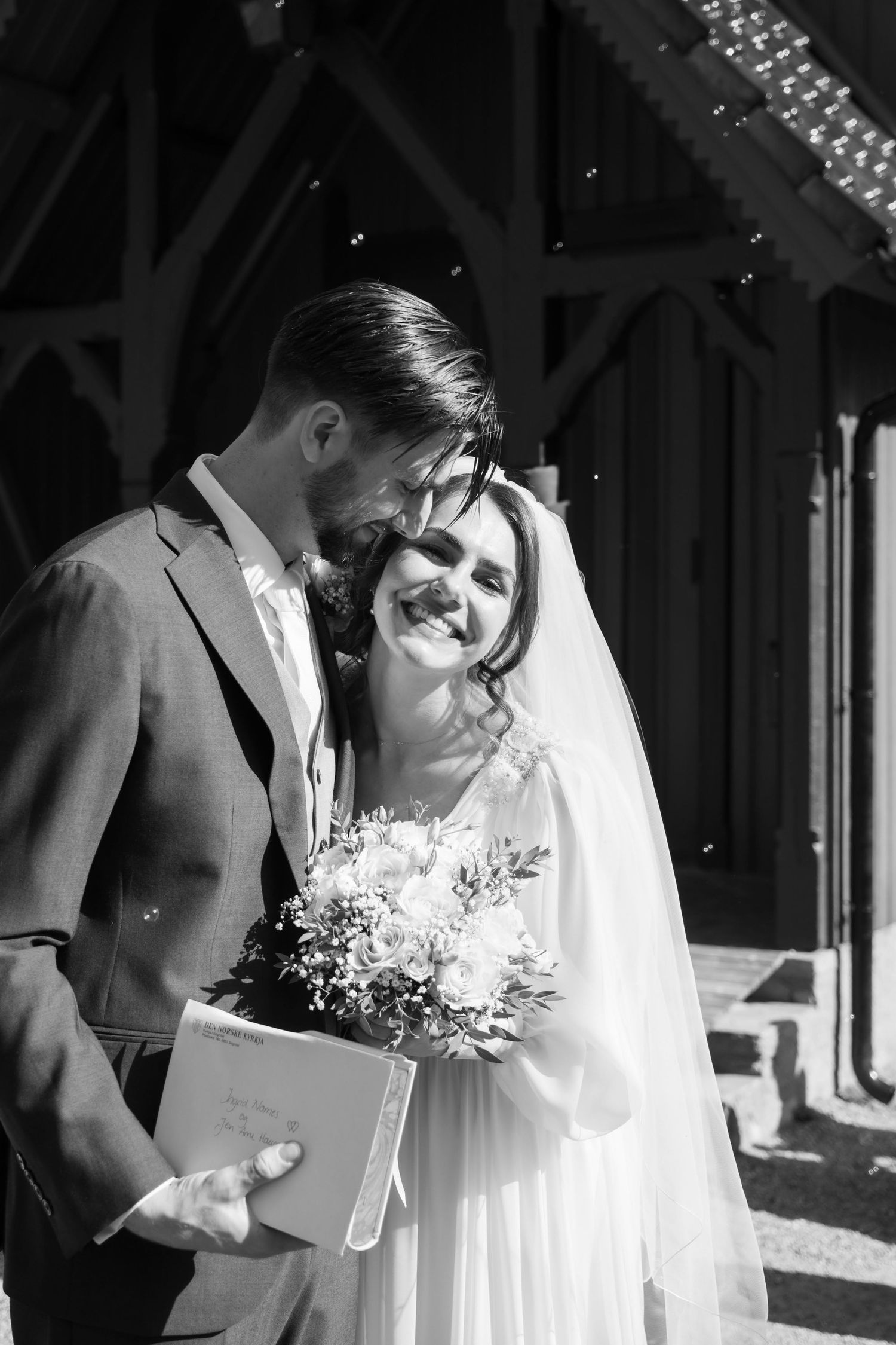 Smiling bride holding bouquet stands with groom outside church in elegant black and white photo.