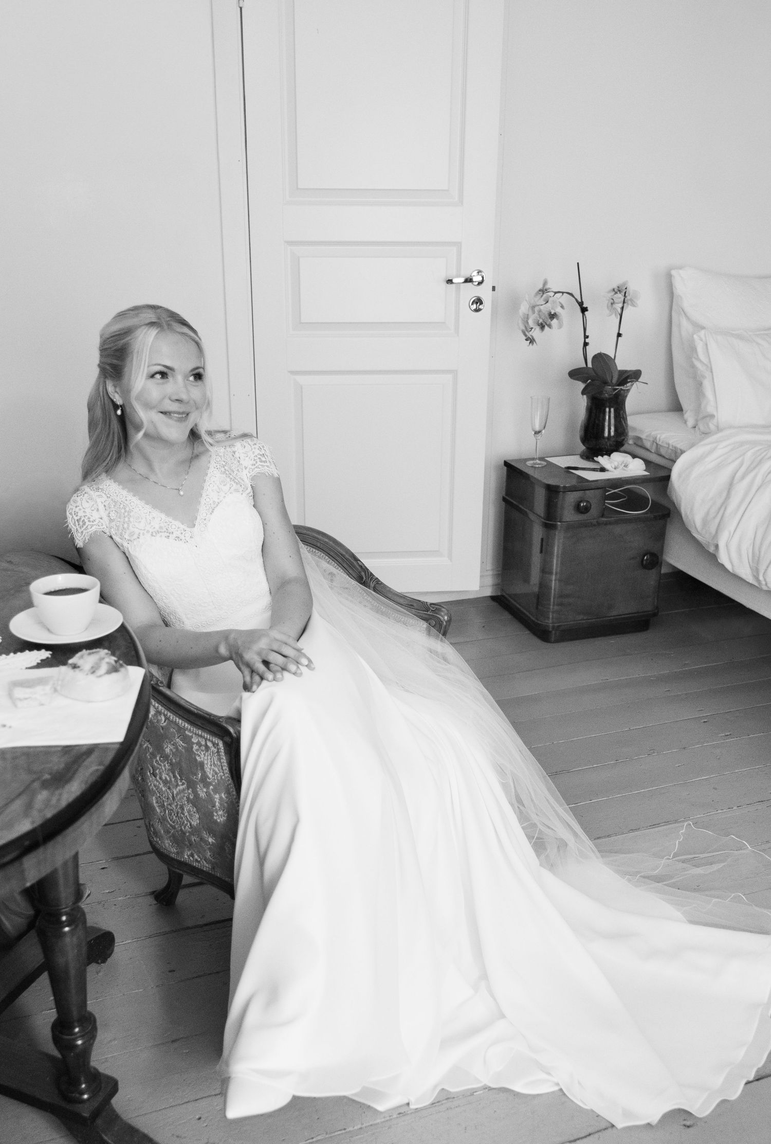 Bride in white lace wedding gown seated at table with coffee cup in elegant room.