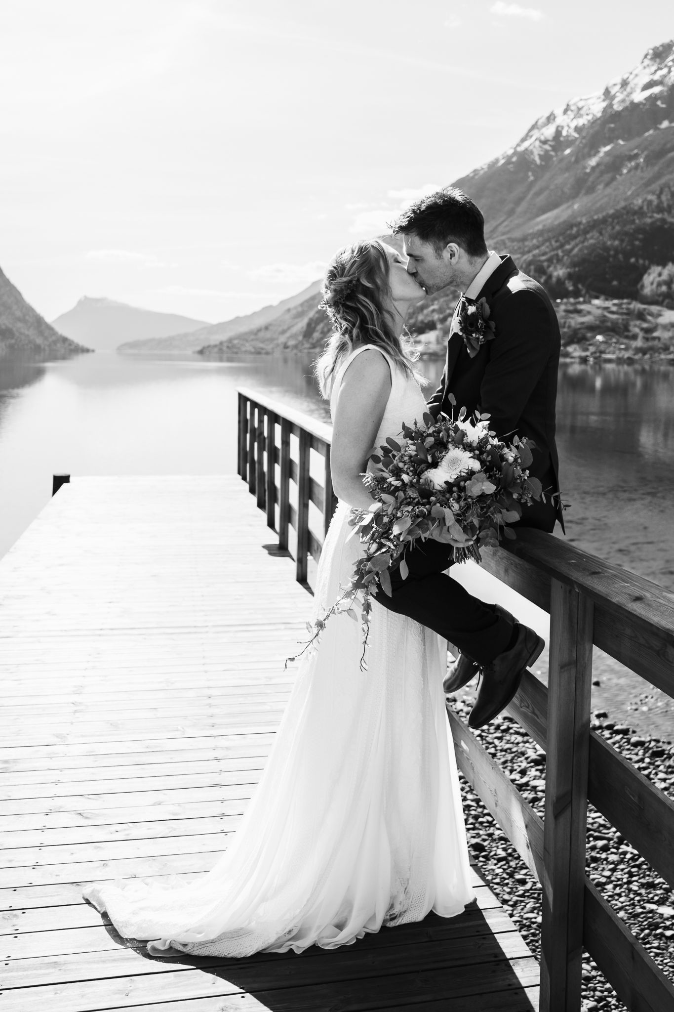 Black and white photo of bride and groom kissing on a wooden dock with mountain lake backdrop.