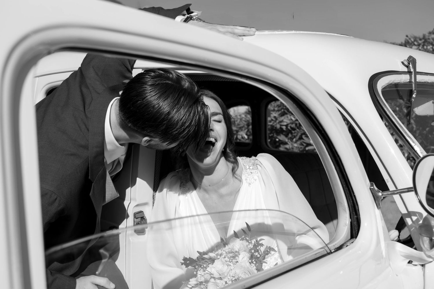 Black and white photo of a bride and groom sharing a kiss through a vintage car window on their wedding day.