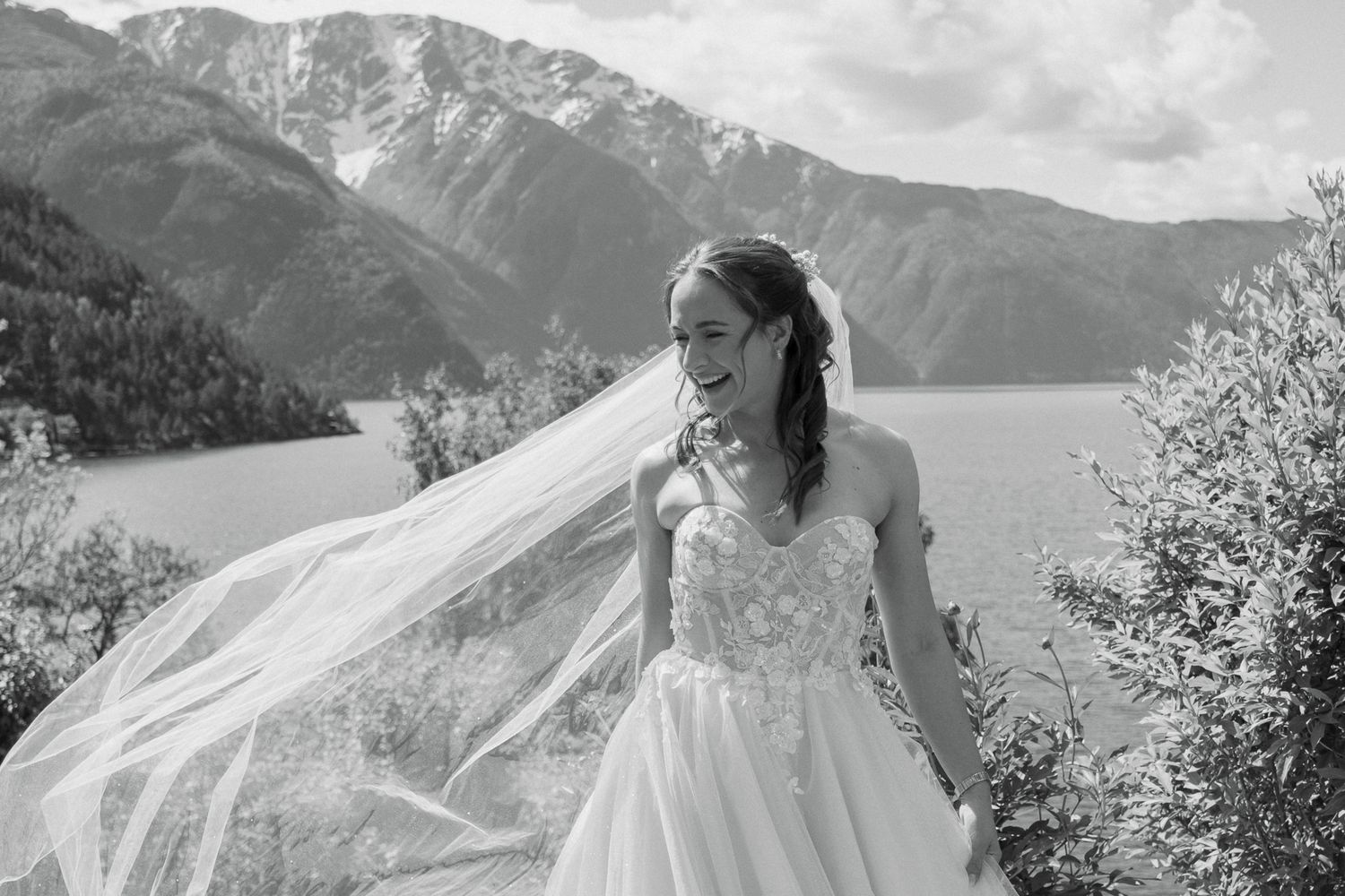 Black and white photo of joyful bride with flowing veil in lace gown against mountain fjord backdrop.