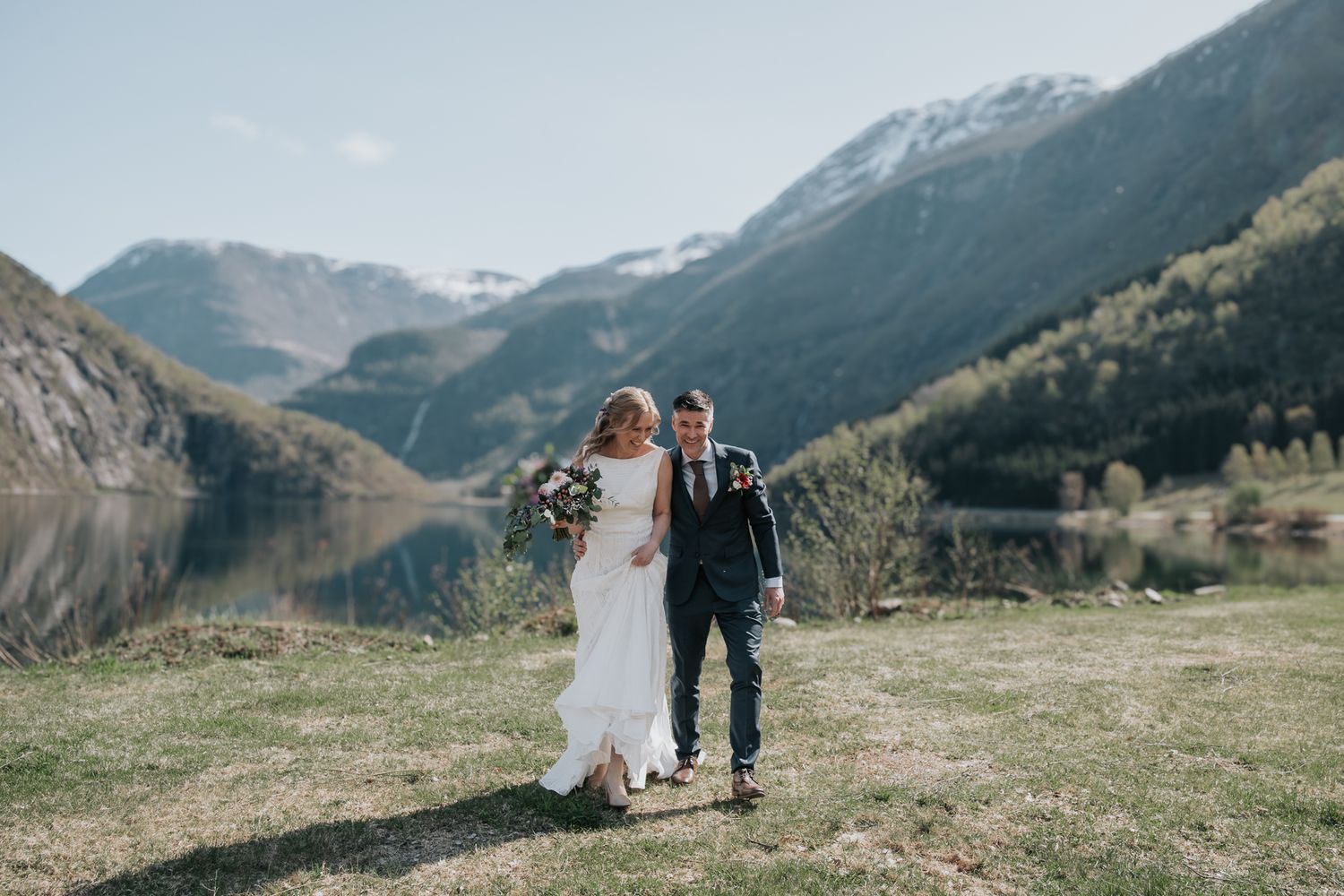 Bride and groom walking together on a grassy lakeside with stunning snow-capped Norwegian mountain scenery.