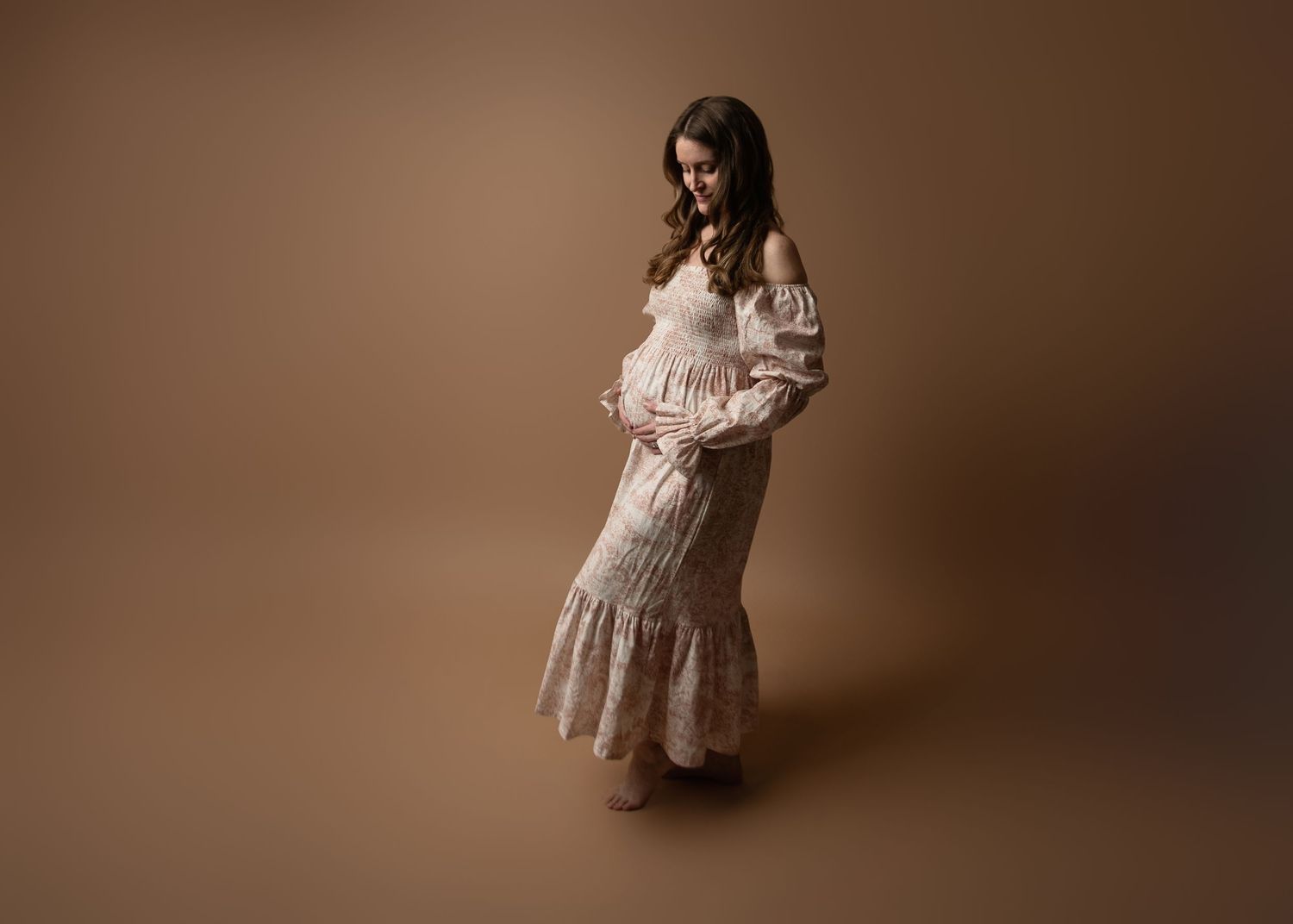 Elegant maternity portraits featuring woman in flowing cream dress against brown backdrop.