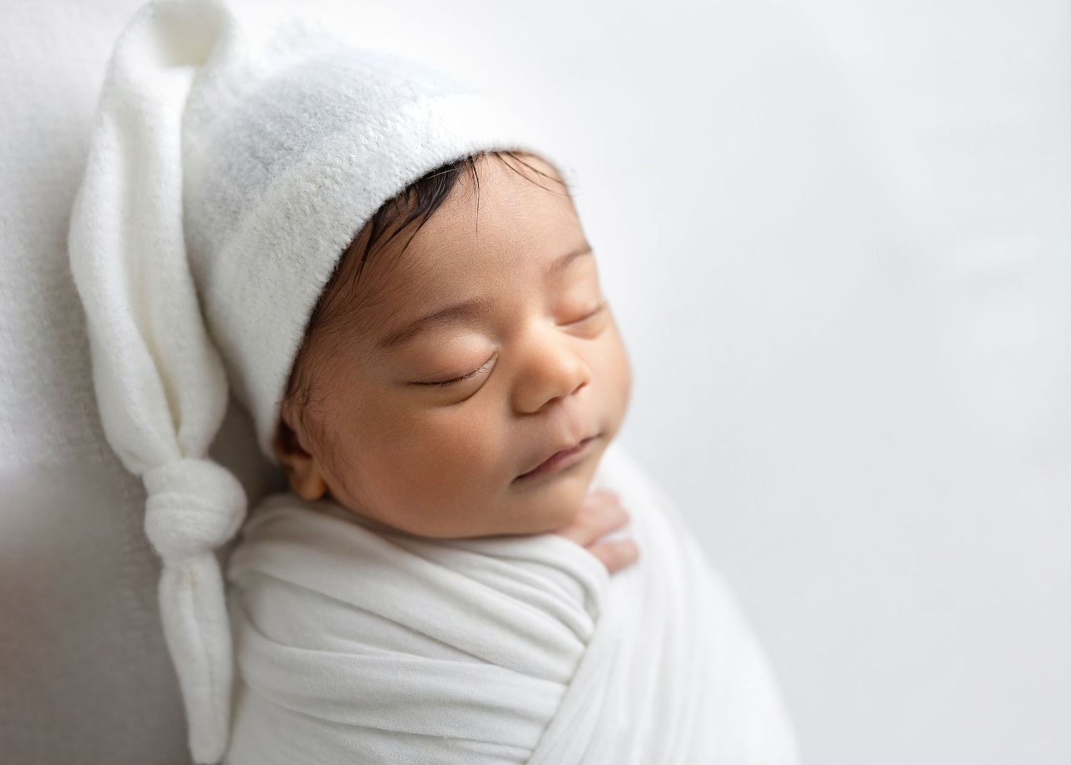 Peaceful newborn baby sleeping wrapped in white swaddle cloth wearing a white knotted sleep cap.
